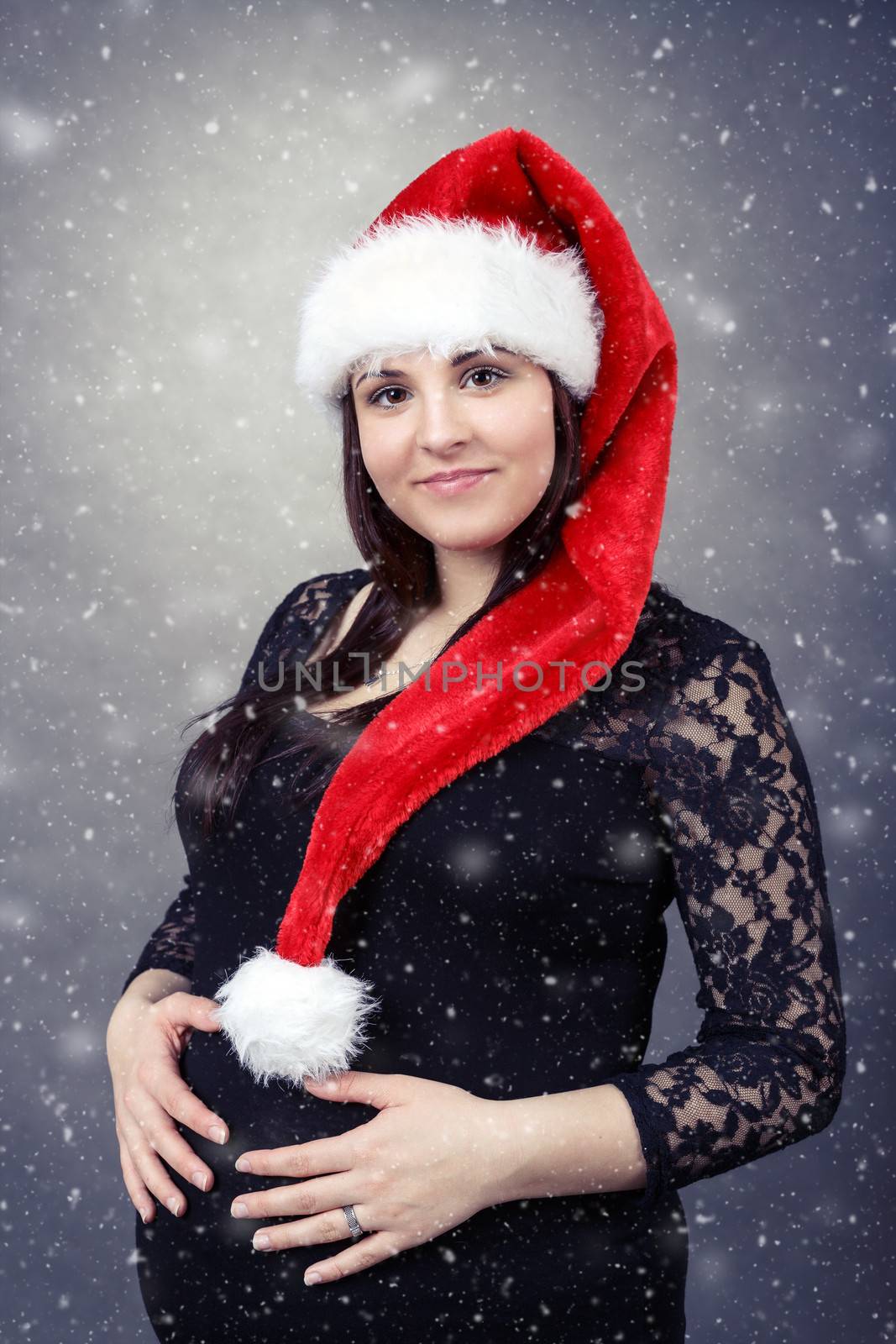 beautiful smiling pregnant young santa woman tenderly holding her tummy with snowing in background