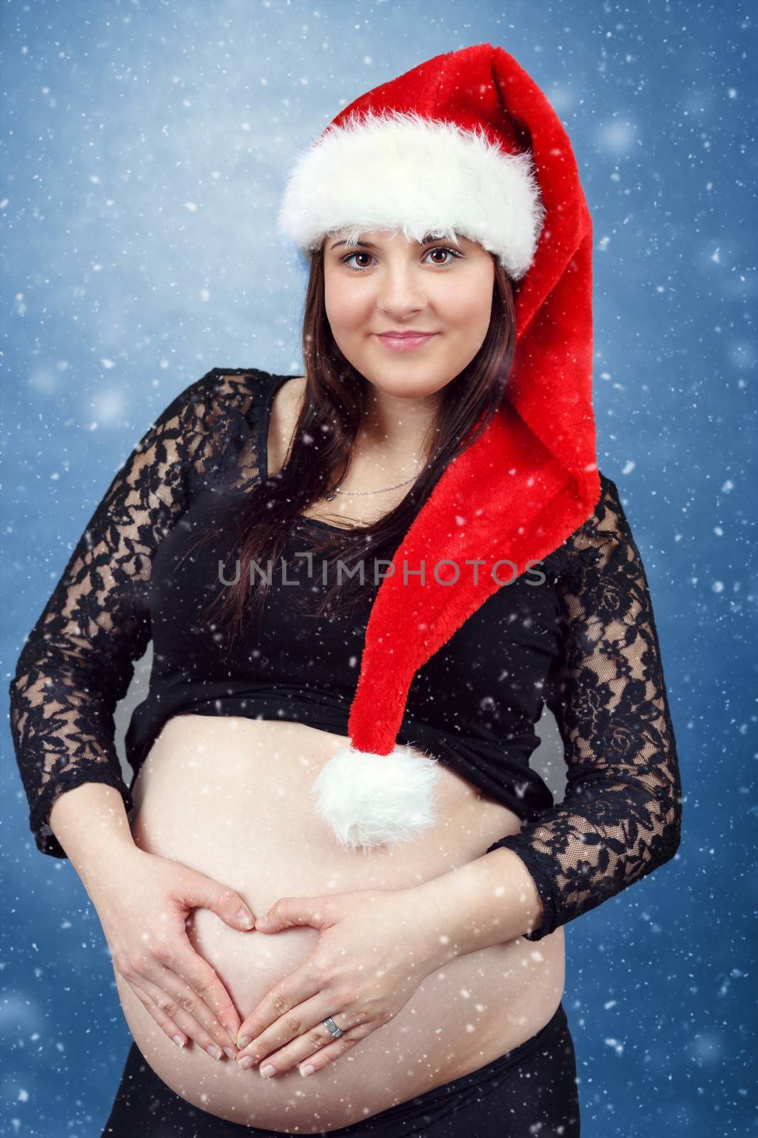 beautiful smiling pregnant young santa woman tenderly holding her tummy, hand in heart shape and with snowing in background