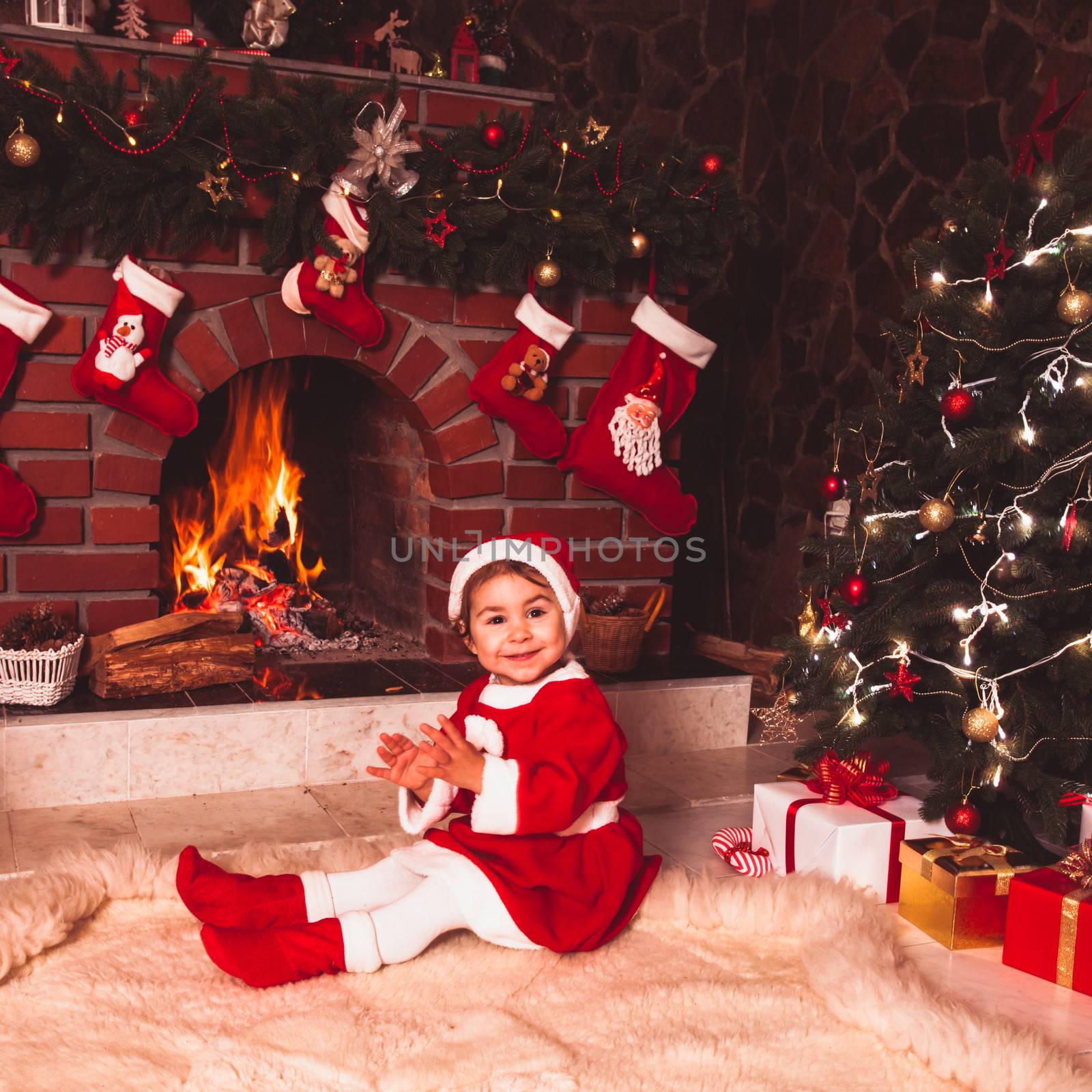 Little girl sitting near fireplace and christmas tree with gift boxes