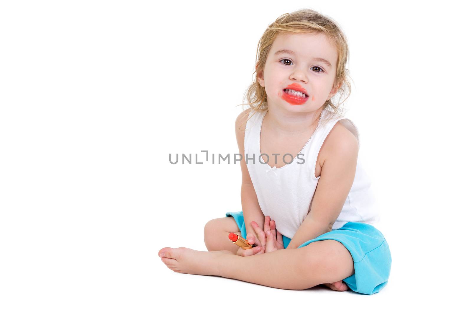 Cute very young girl practising to be a fashion model sitting on the floor grinning at the camera with her mouth smeared with bright red lipstick which she is holding in her hand, on white