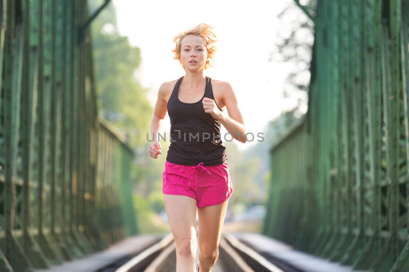 Athlete running on railaway tracks bridge in morning sunrise training for marathon and fitness. Healthy sporty caucasian woman exercising in urban environment before going to work; Active urban lifestyle.