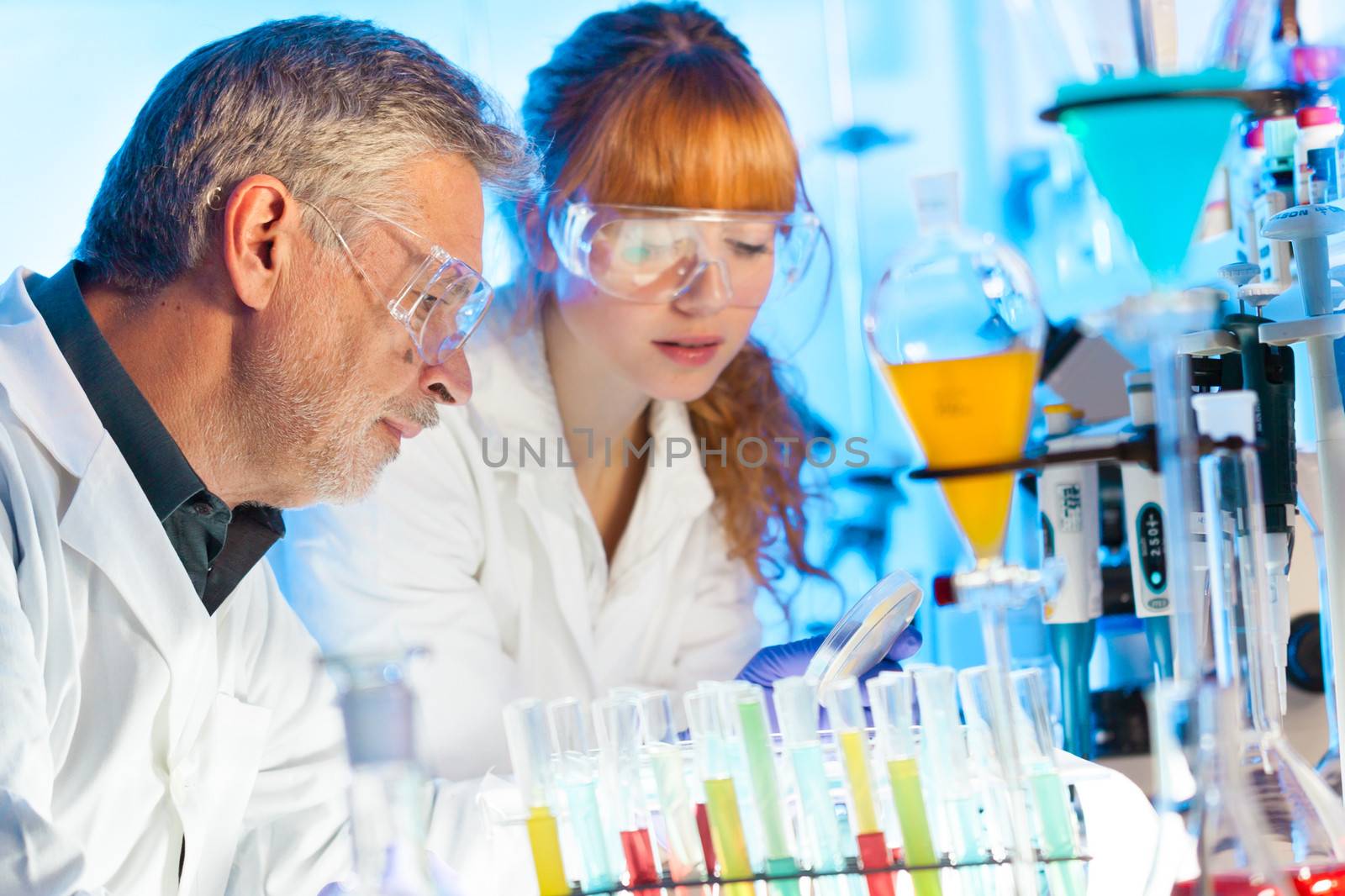 Attractive young female scientist and her senior male supervisor observing color shift of a red liquid in the glass tube in the life science research laboratory (biochemistry, genetics, forensics, microbiology..)