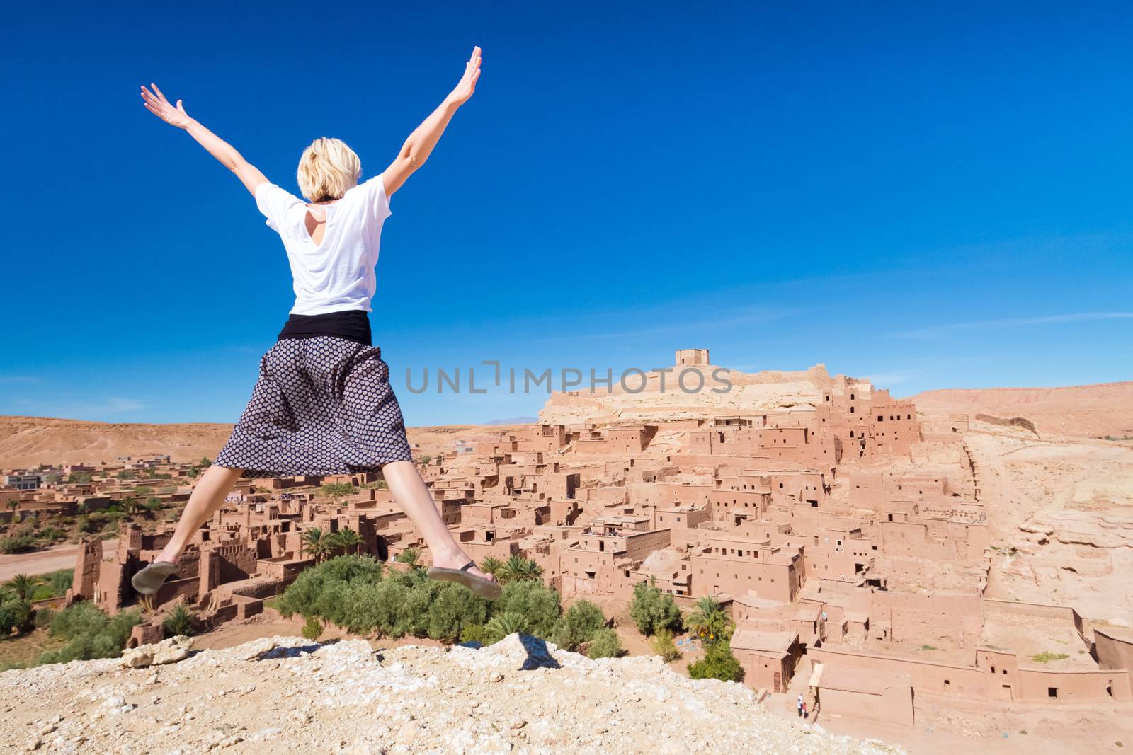 Beautiful cheerful adventurous woman jumping in front of Ait Benhaddou, fortified city, kasbah or ksar, along the former caravan route between Sahara and Marrakesh in present day Morocco.