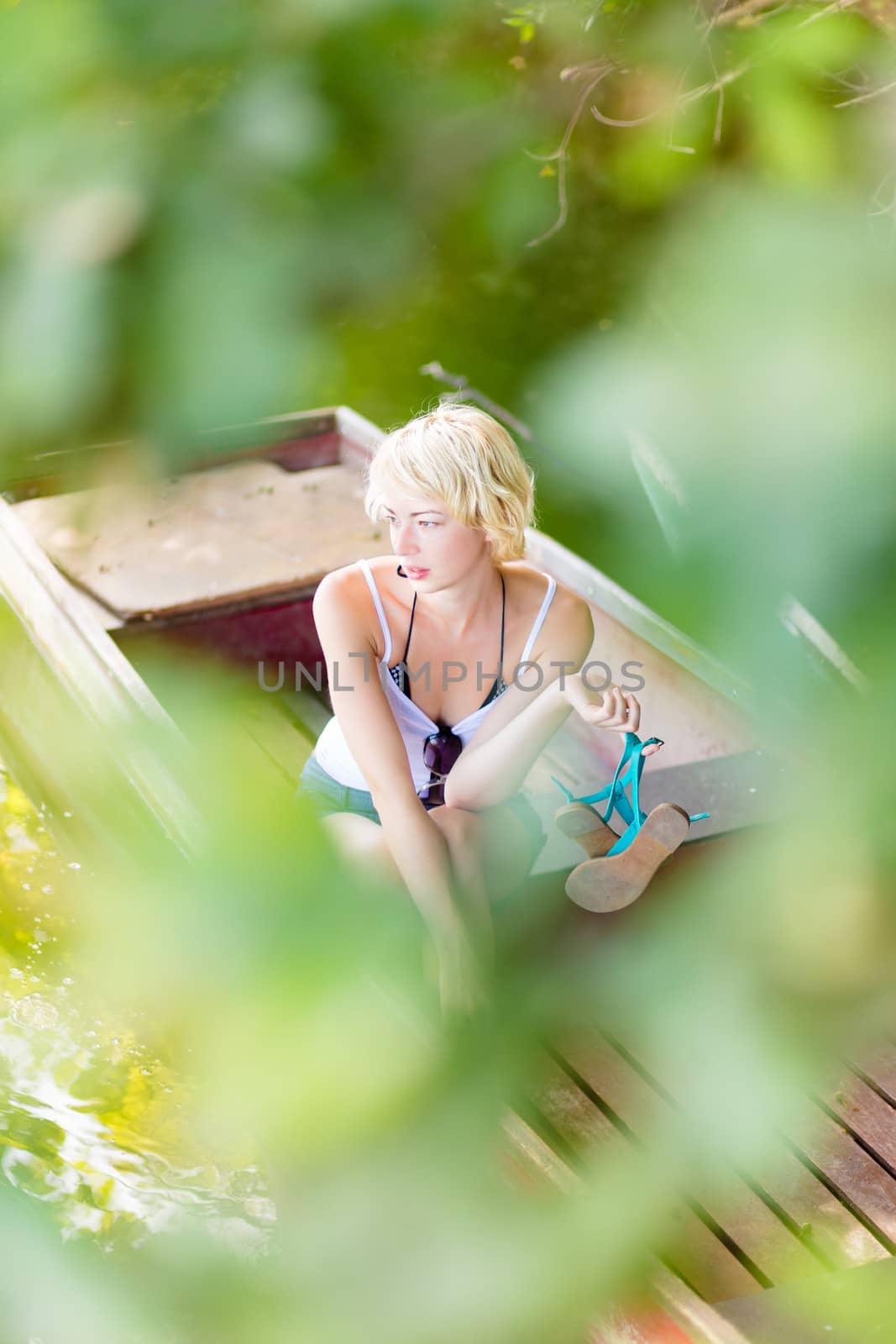 Thoughtful young blonde woman enjoying the sunny summer day on a vintage wooden boat on a lake in pure natural environment  being secretly observed through the branches.