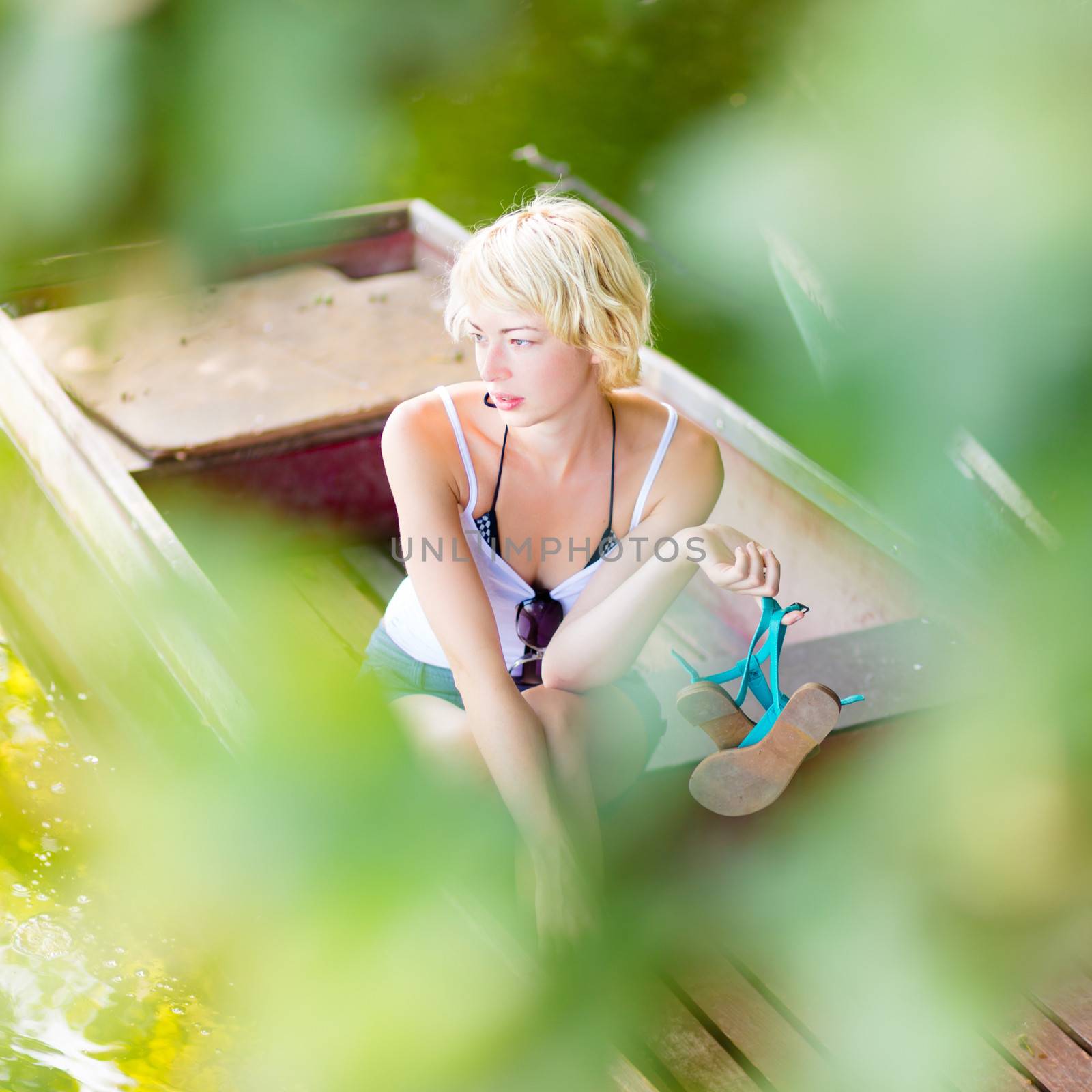 Thoughtful young blonde woman enjoying the sunny summer day on a vintage wooden boat on a lake in pure natural environment  being secretly observed through the branches.