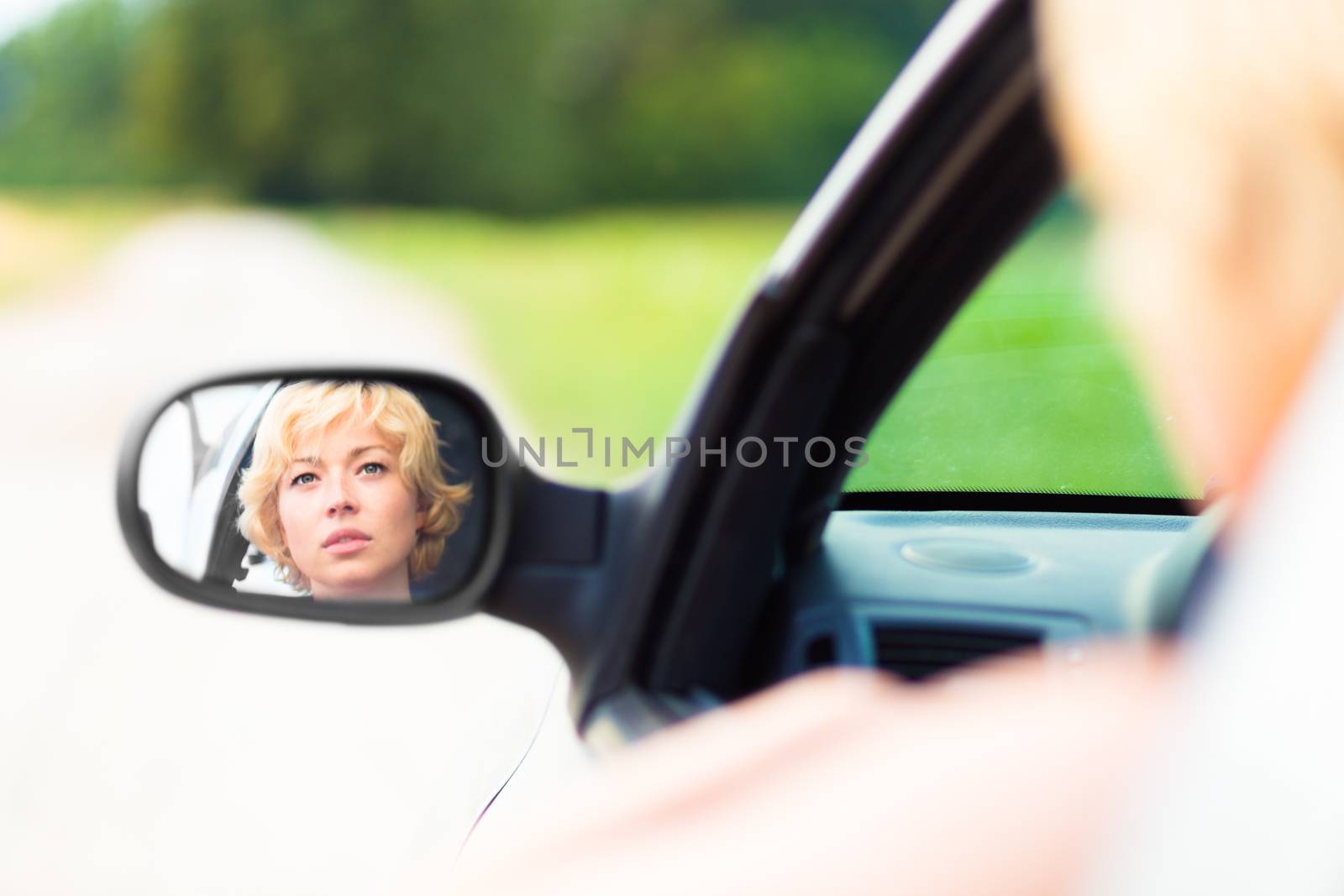 Lady driving a car in the countryside. Drivers face can be seen in the side mirror.