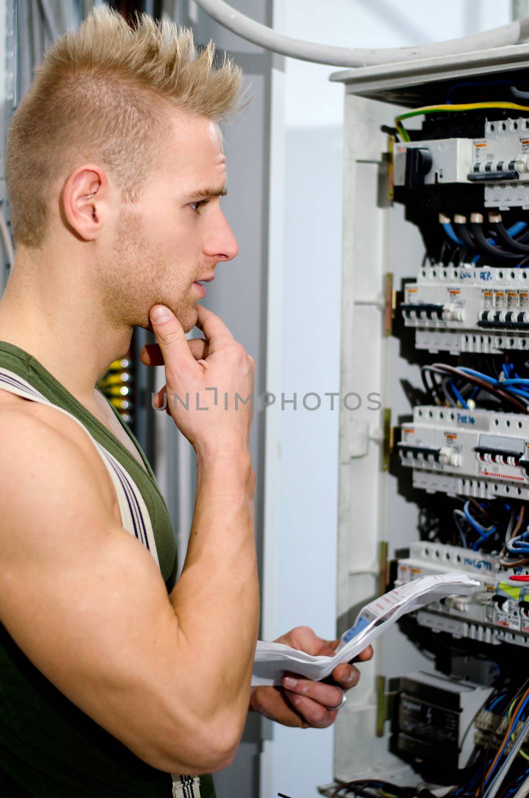 Confused male electrician in front of power panel, holding his chin puzzled