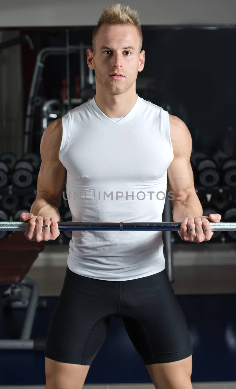 Handsome young man training biceps lifting barbell, standing in a gym
