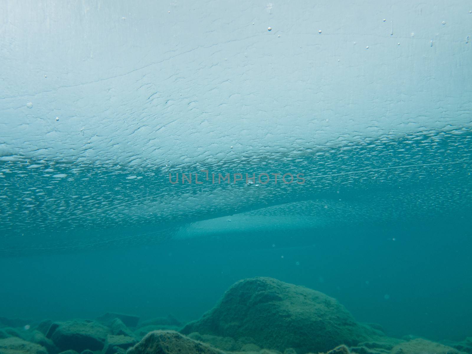 Underwater shot of solid ice sheet covering clear shallow lake water with rocky bottom