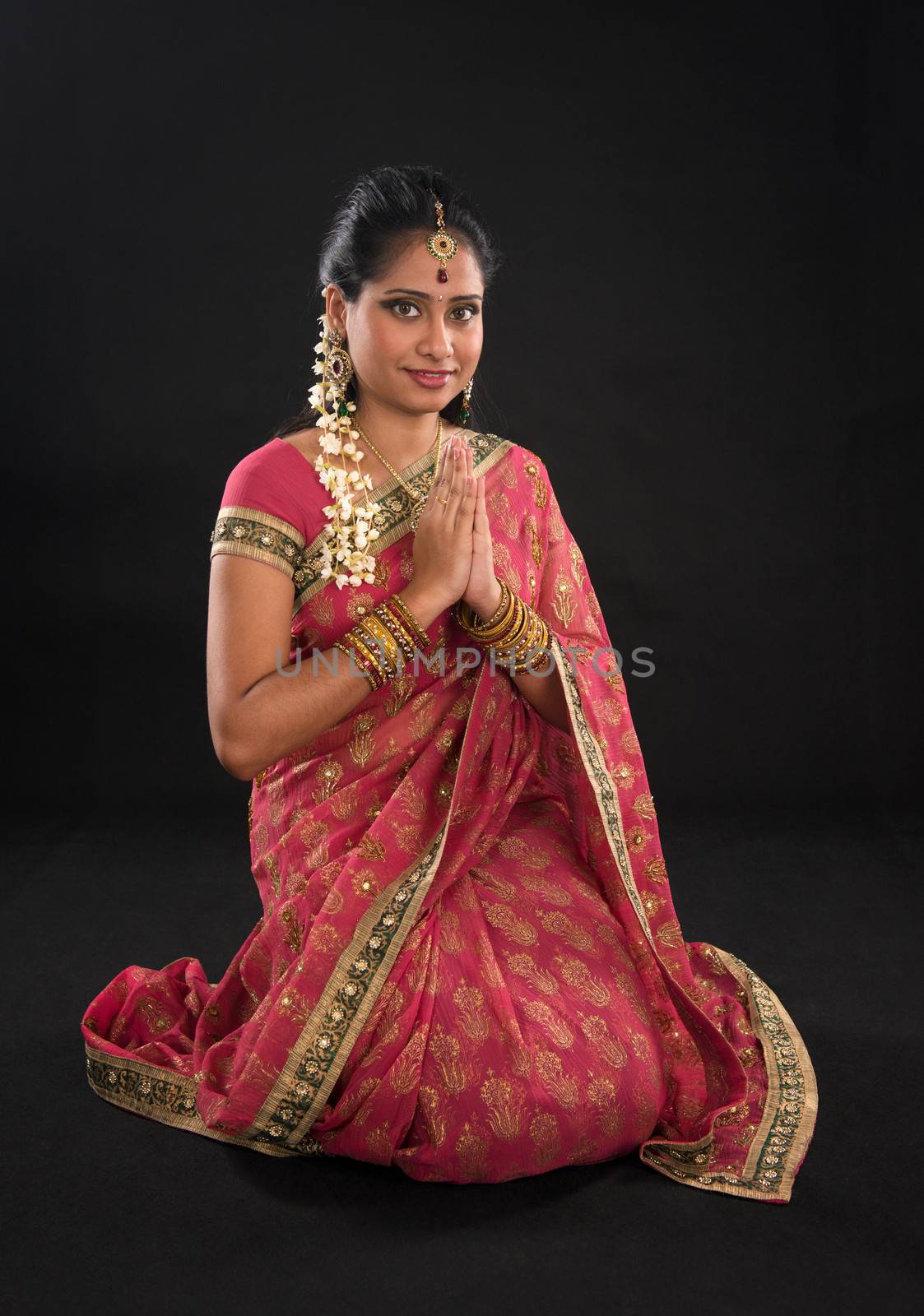 Indian girl in a greeting pose, traditional sari costume, full length kneeling on floor isolated on black background
