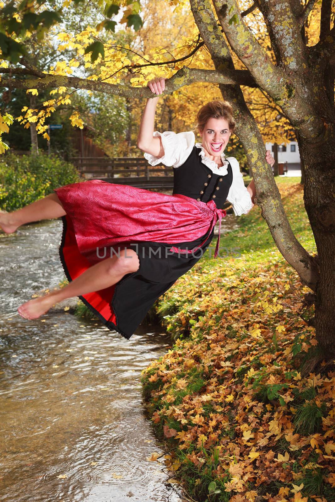 Playful barefoot young woman in a traditional dirndl swinging on the branches an autumn tree over a stream laughing with happiness