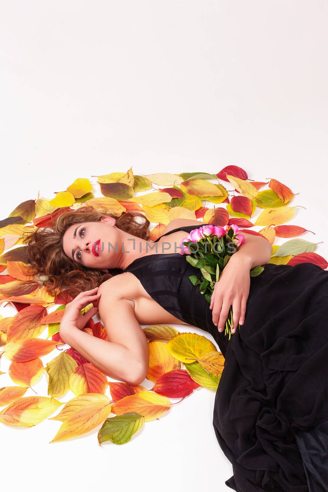 Beautiful sexy woman in an elegant simple black dress lying on a carpet of yellow and red autumn leaves with a bouquet of roses dreaming of a loved one