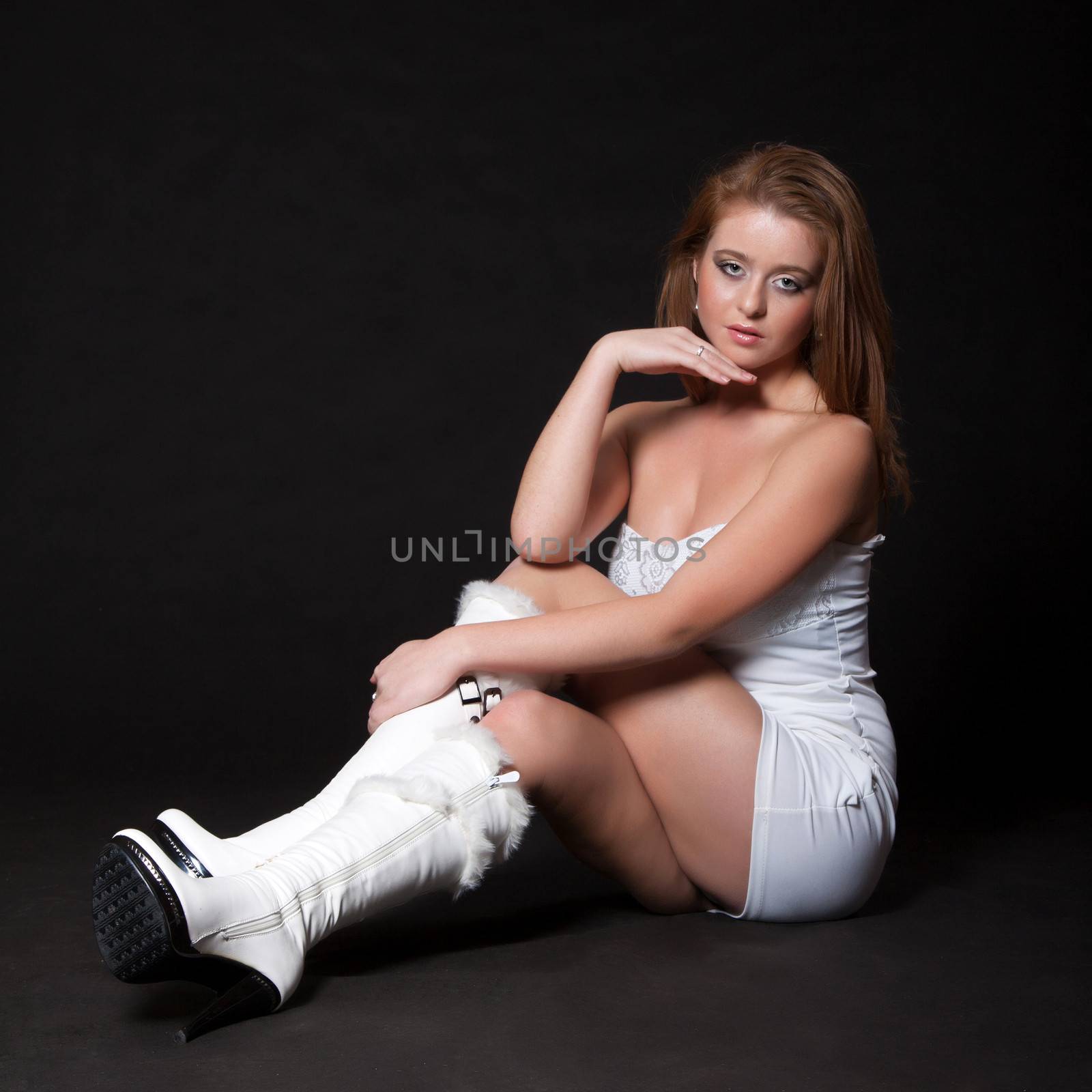 Young fashion model in a white mini dress sitting on the floor and looking up with a dreamy look on a black background