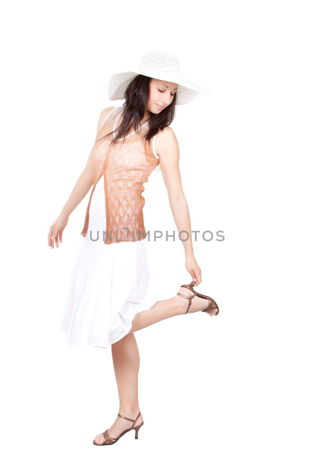 Brunette woman in white sun hat in a white dress, on white background, looking at the raised leg heel