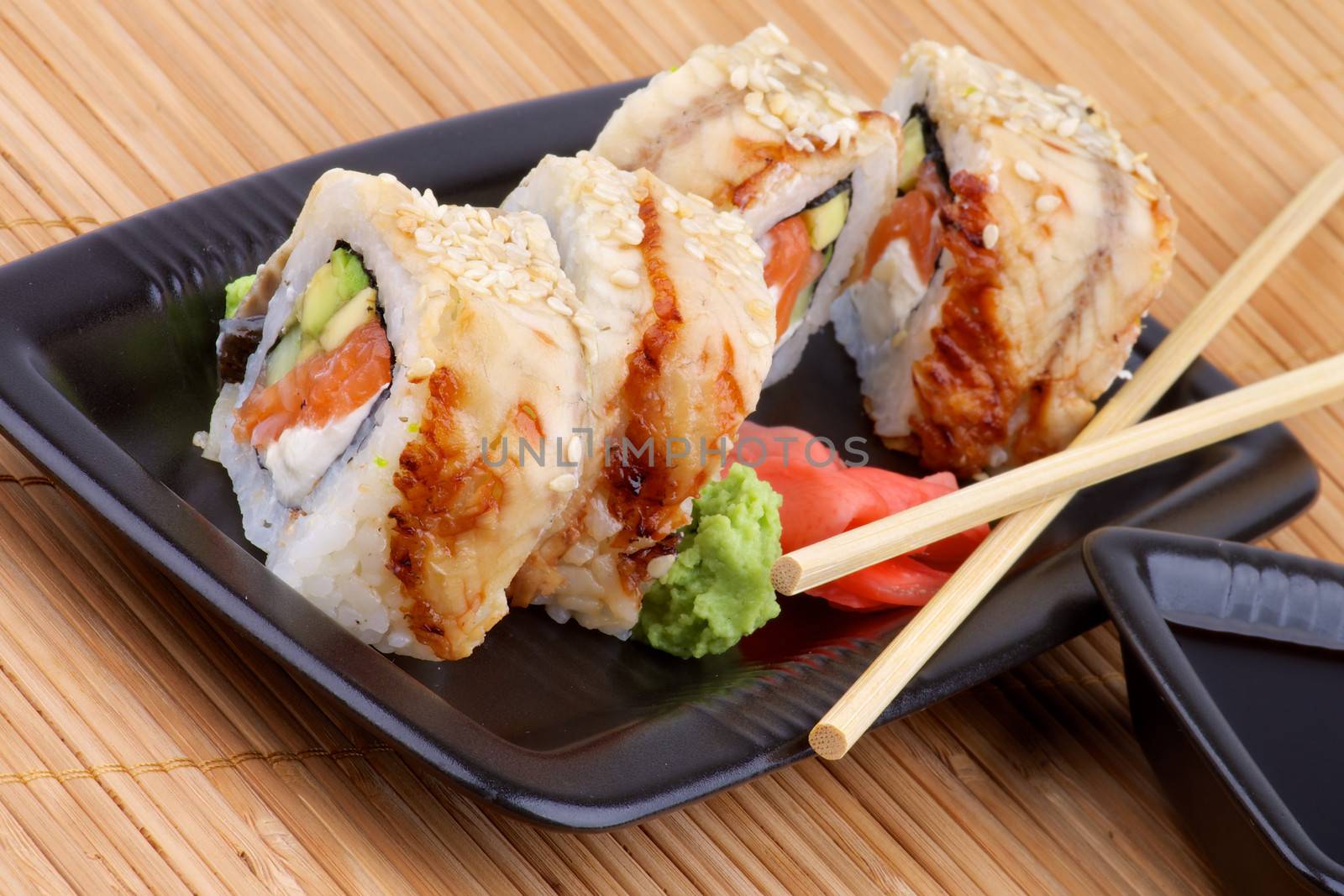 Arrangement of Unagi Sushi Rolls with Wasabi, Marinated Ginger and Soy Sauce in Black Square Shape Bowls with Pair of Chopsticks on Straw mat background