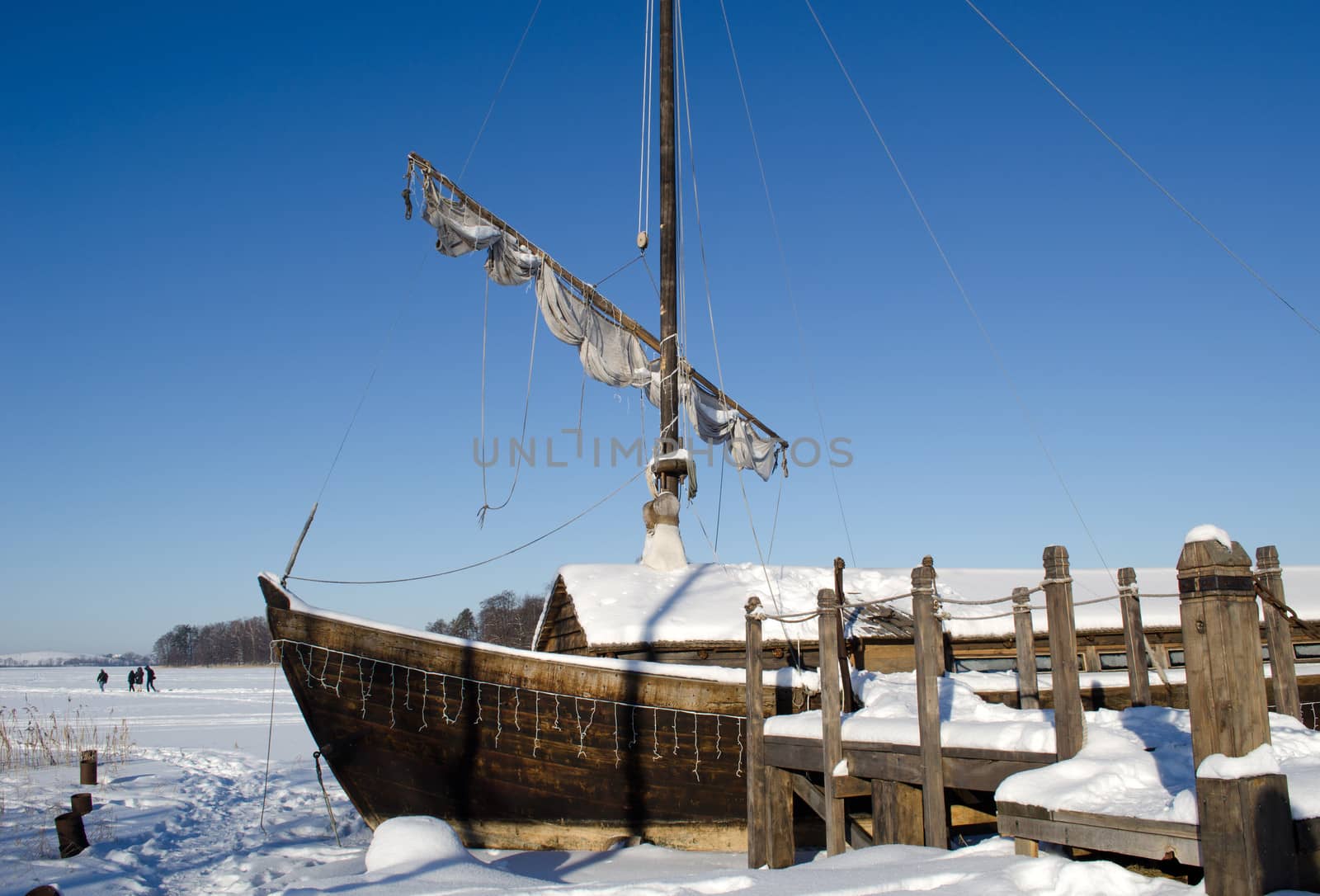 retro wooden ship frozen in lake ice near pier and sail on background of blue sky. people recreate in nature.