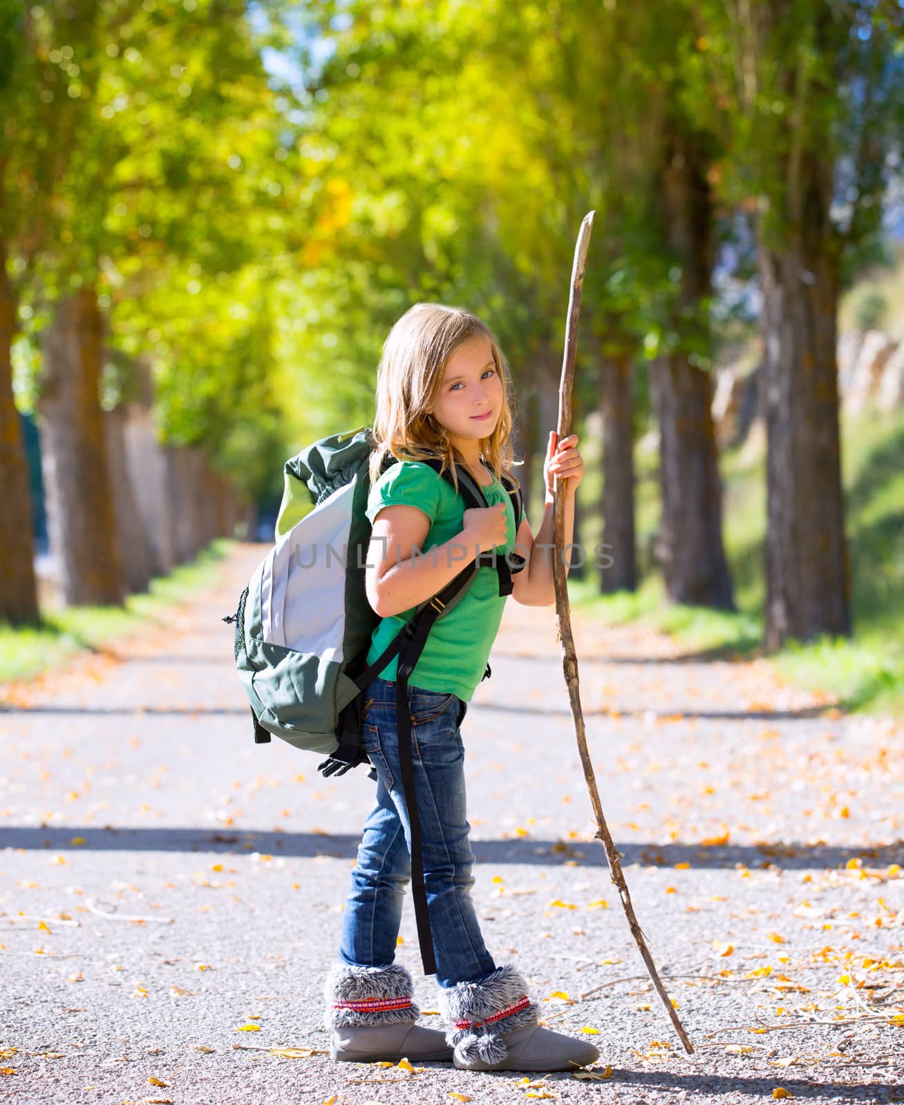 Blond explorer kid girl walking with backpack hiking in autumn trees track holding stick