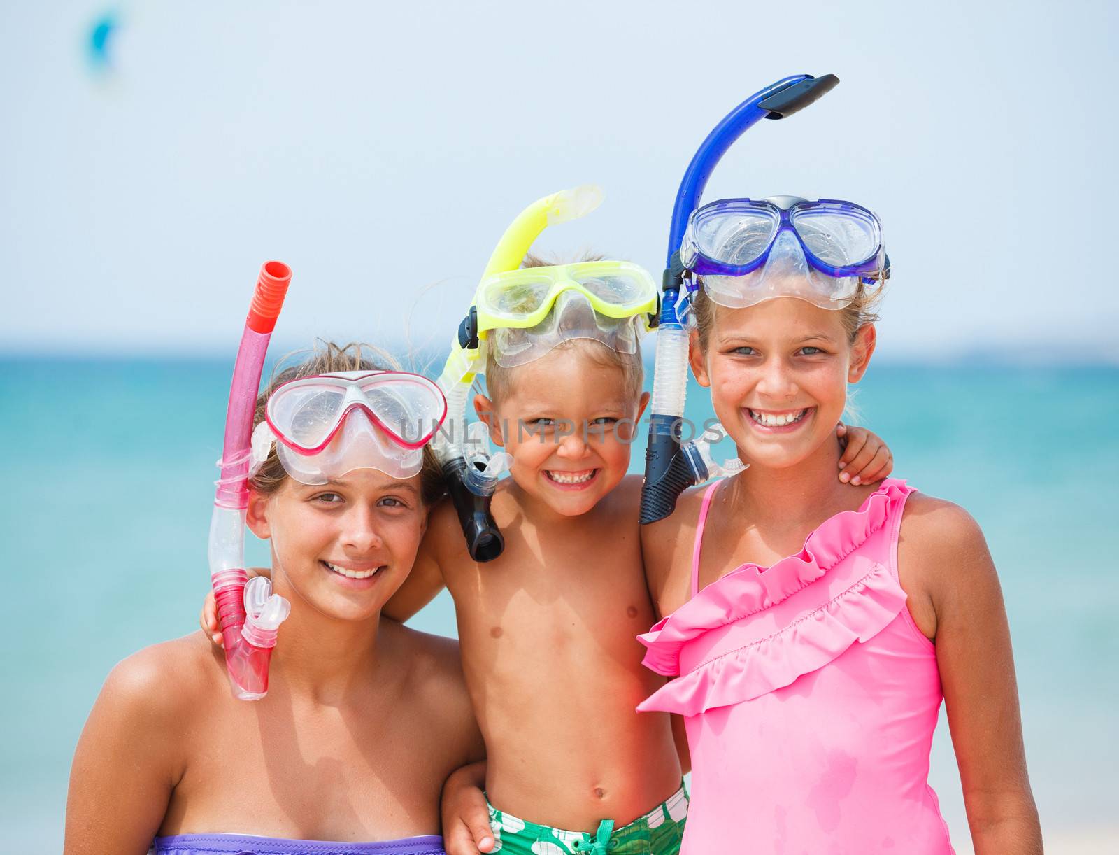 Three happy children on beach with colorful face masks and snorkels, sea in background.