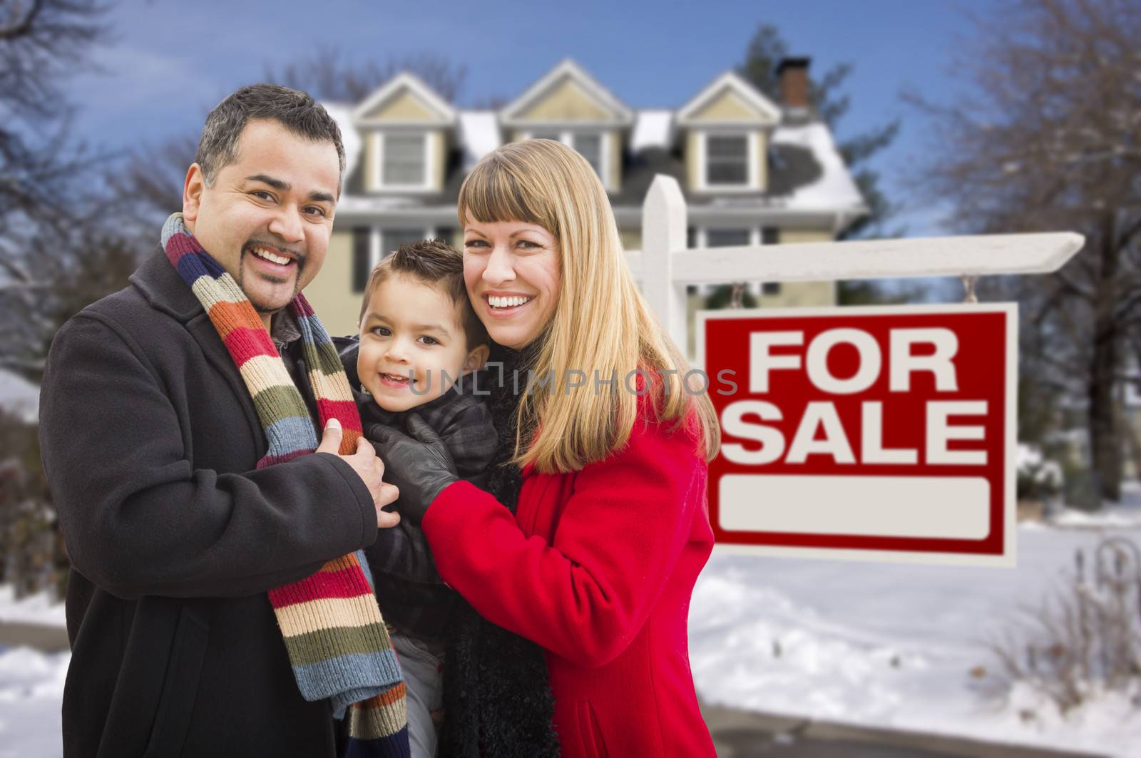 Warmly Dressed Young Mixed Race Family in Front of Home For Sale Real Estate Sign and House with Snow On The Ground.