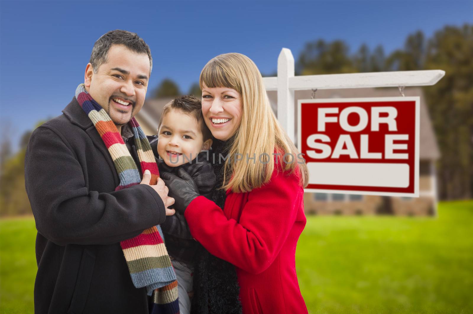 Warmly Dressed Young Mixed Race Family in Front of Home For Sale Real Estate Sign and House.