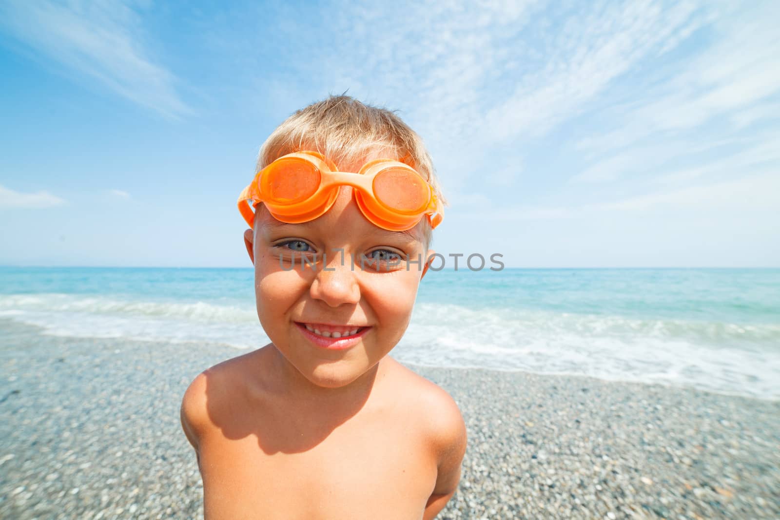 Playful boy in swimming goggles on the beach with sea on background