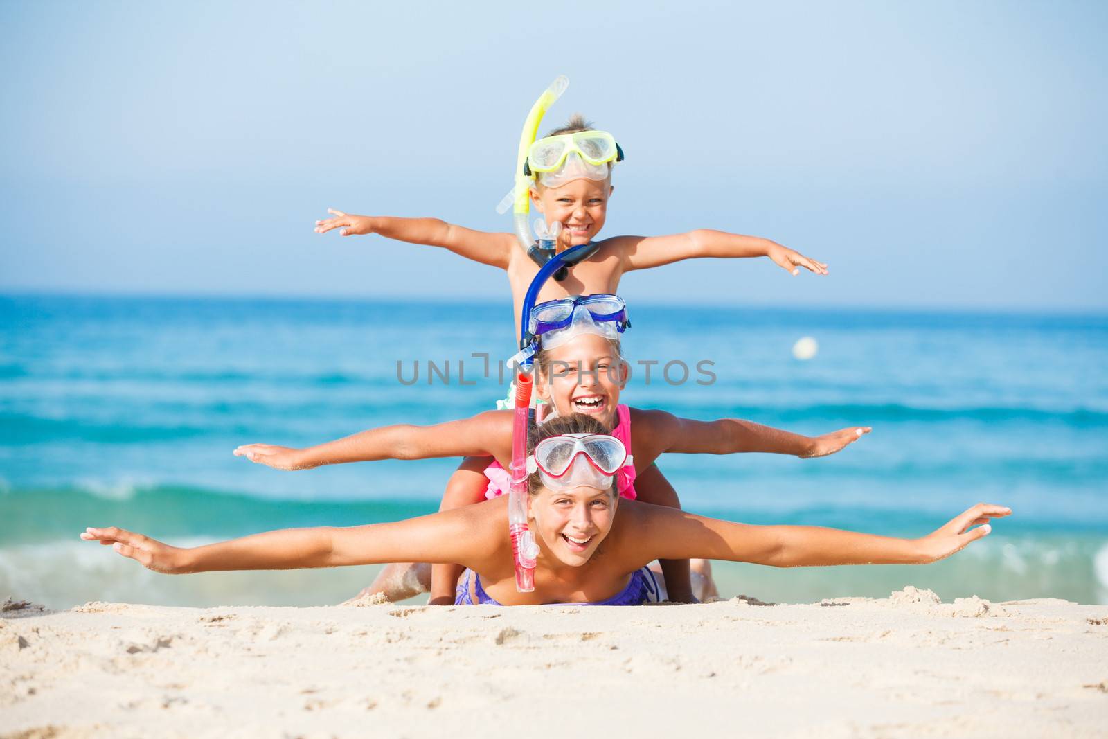 Three happy children on beach with colorful face masks and snorkels, sea in background.