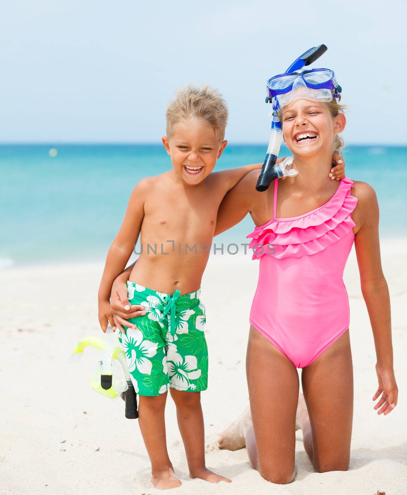 Two happy children on beach with colorful face masks and snorkels, sea in background.