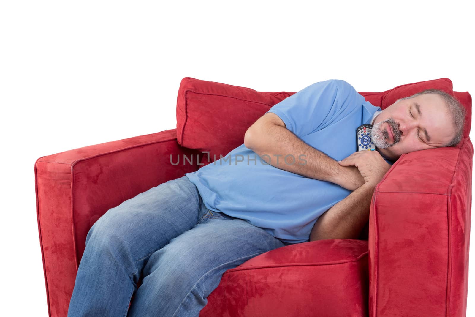 Tired middle-aged man fallen asleep while watching television with the remote control in his hand and his head resting on the arm of a comfortable red chair, isolated on white
