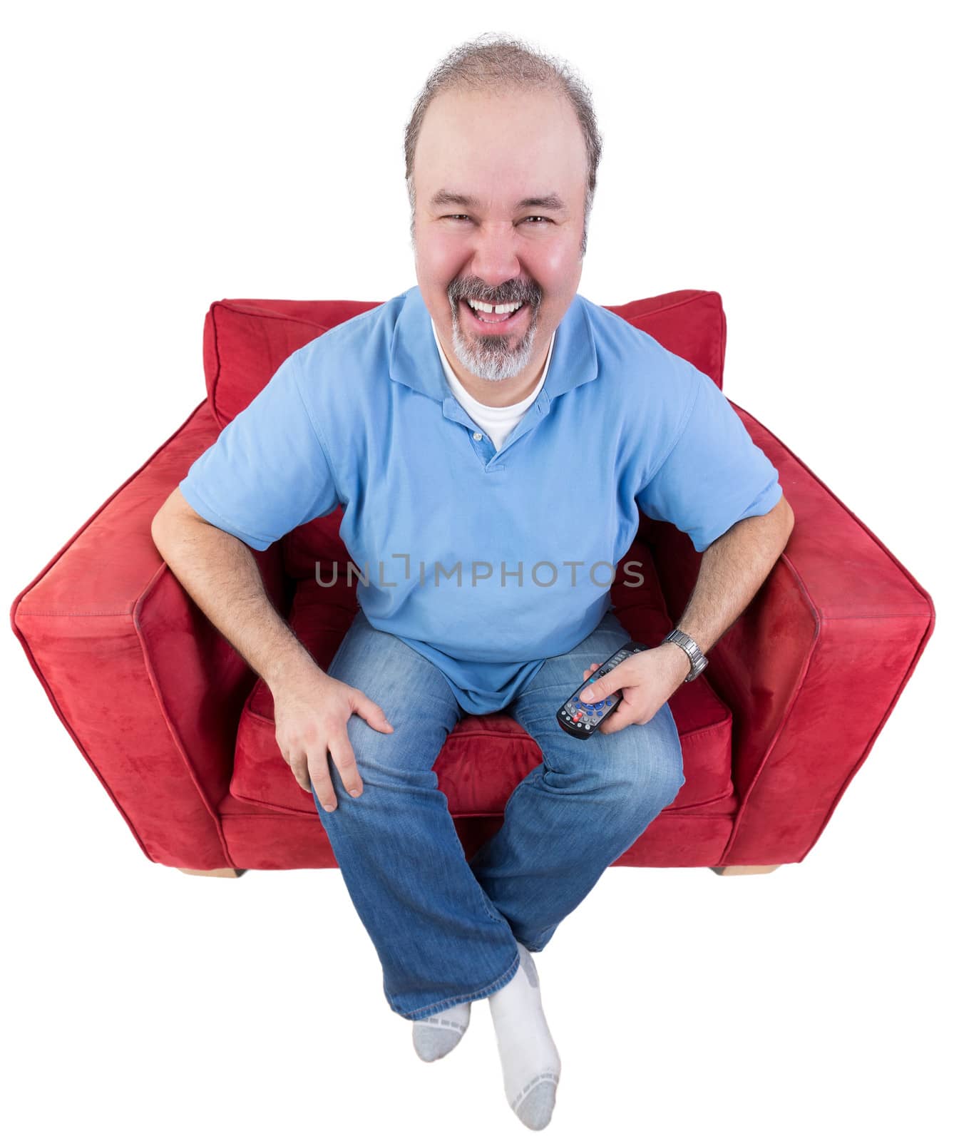 Middle-aged bearded man laughing as he watches television sitting in a comfortable armchair with the remote control in his hand