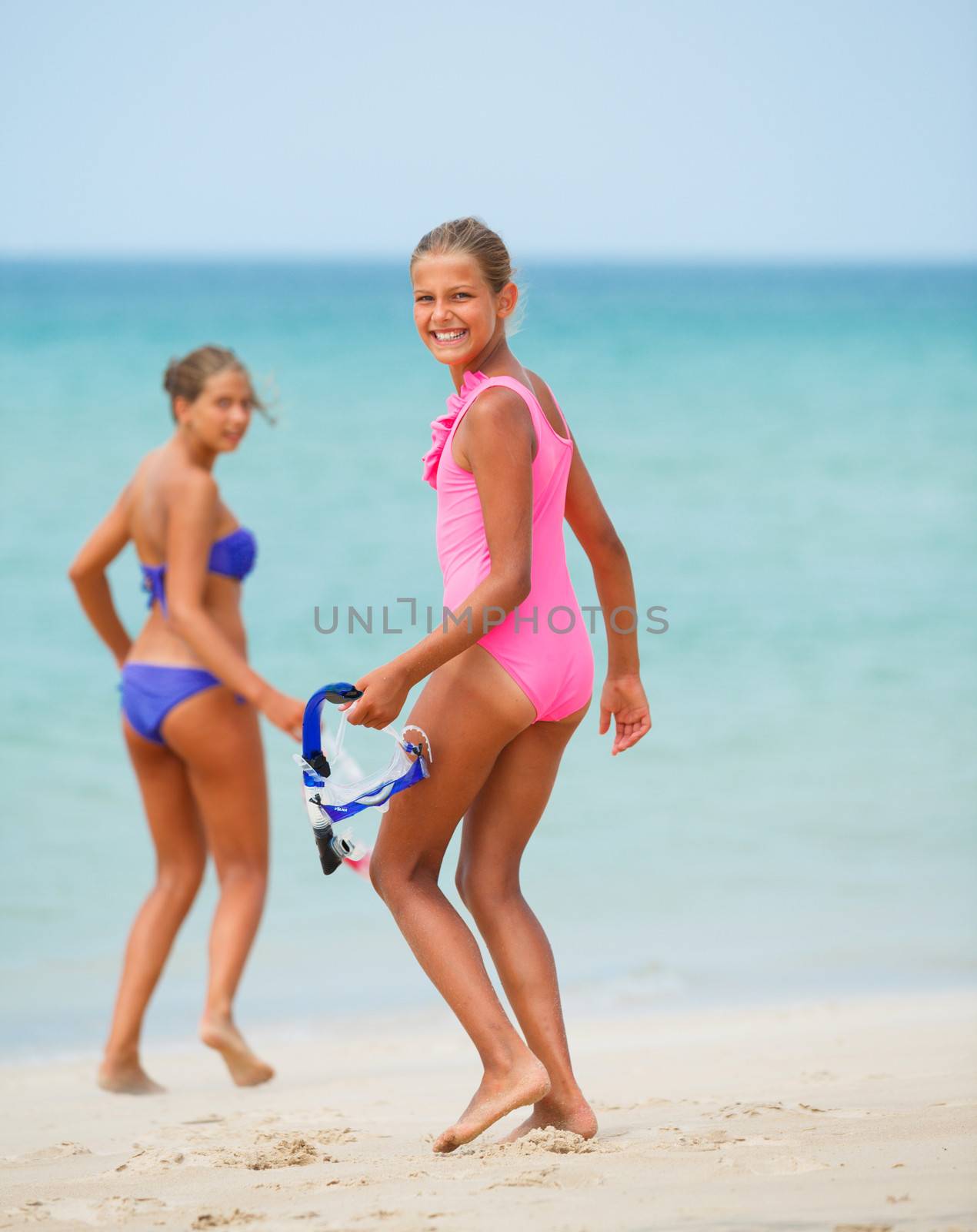 Back view of two happy girls on beach with colorful face masks and snorkels, sea in background.