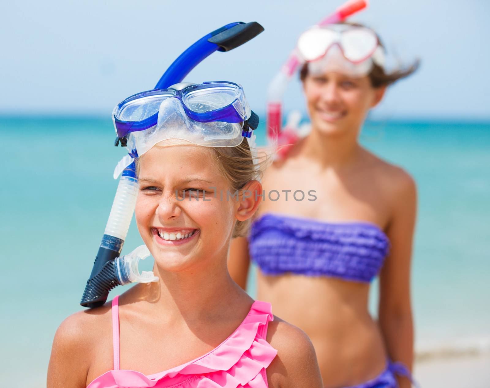 Closeup portrait of two happy girls on beach with colorful face masks and snorkels, sea in background.