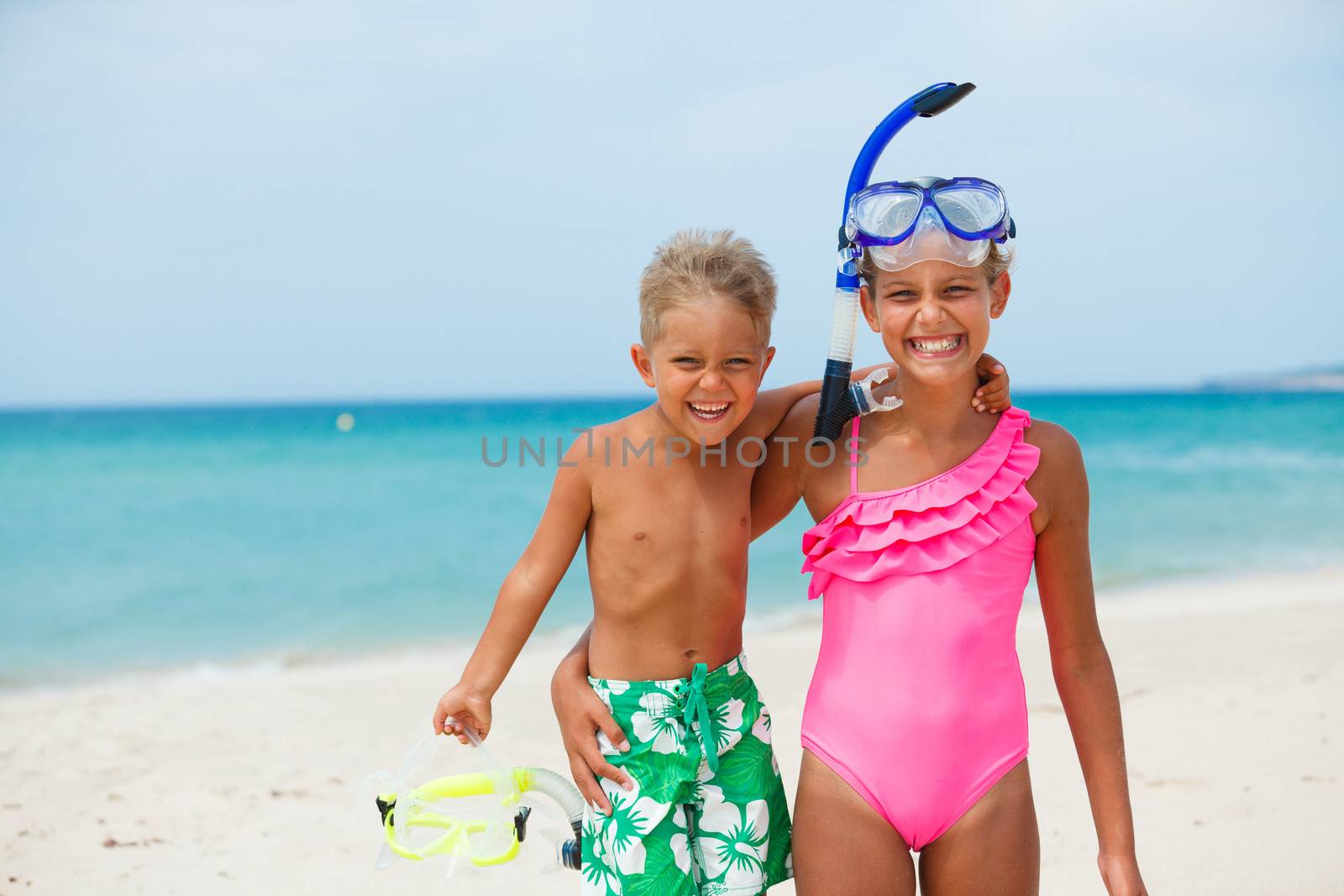 Two happy children on beach with colorful face masks and snorkels, sea in background.