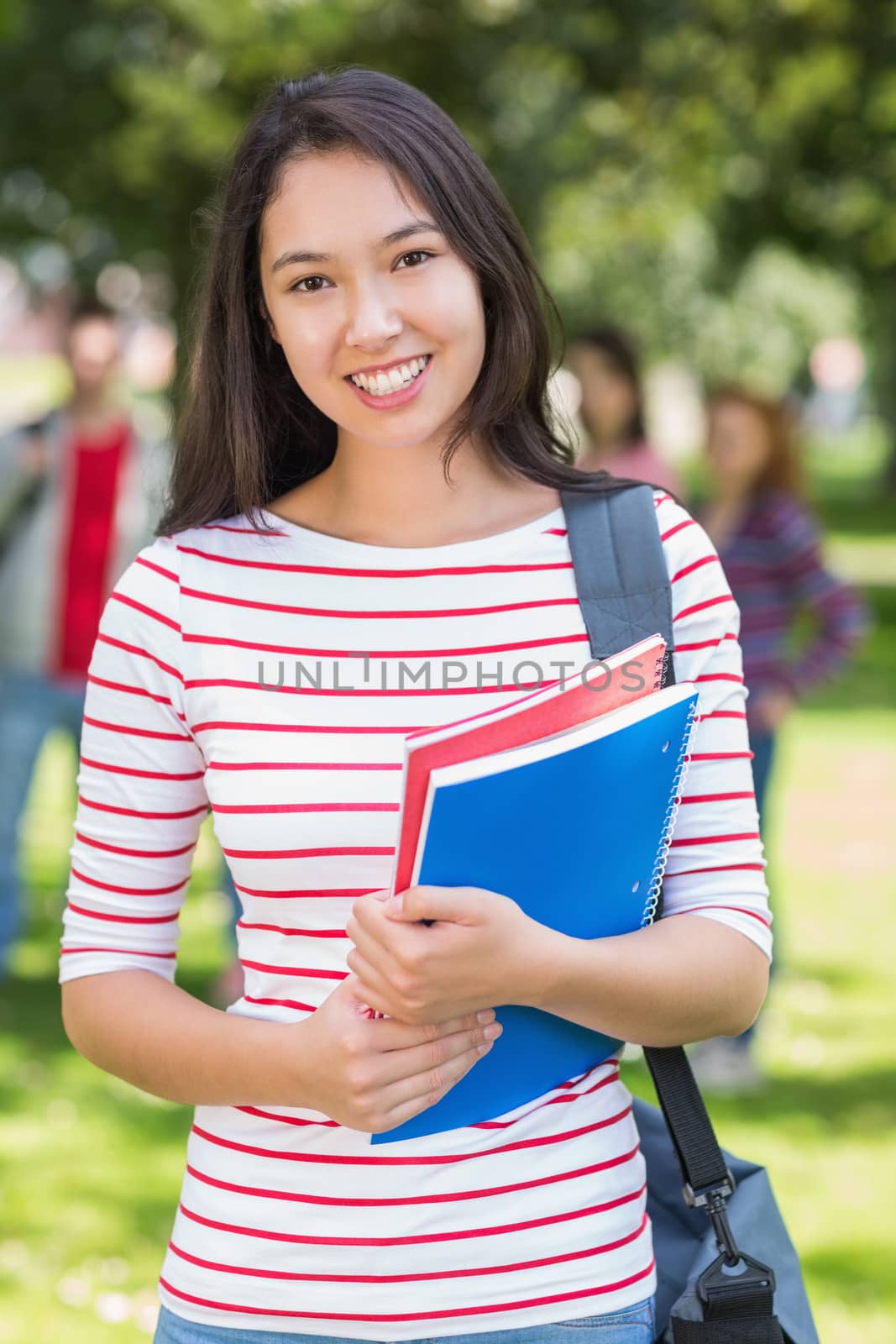 College girl holding books with blurred students in park by Wavebreakmedia