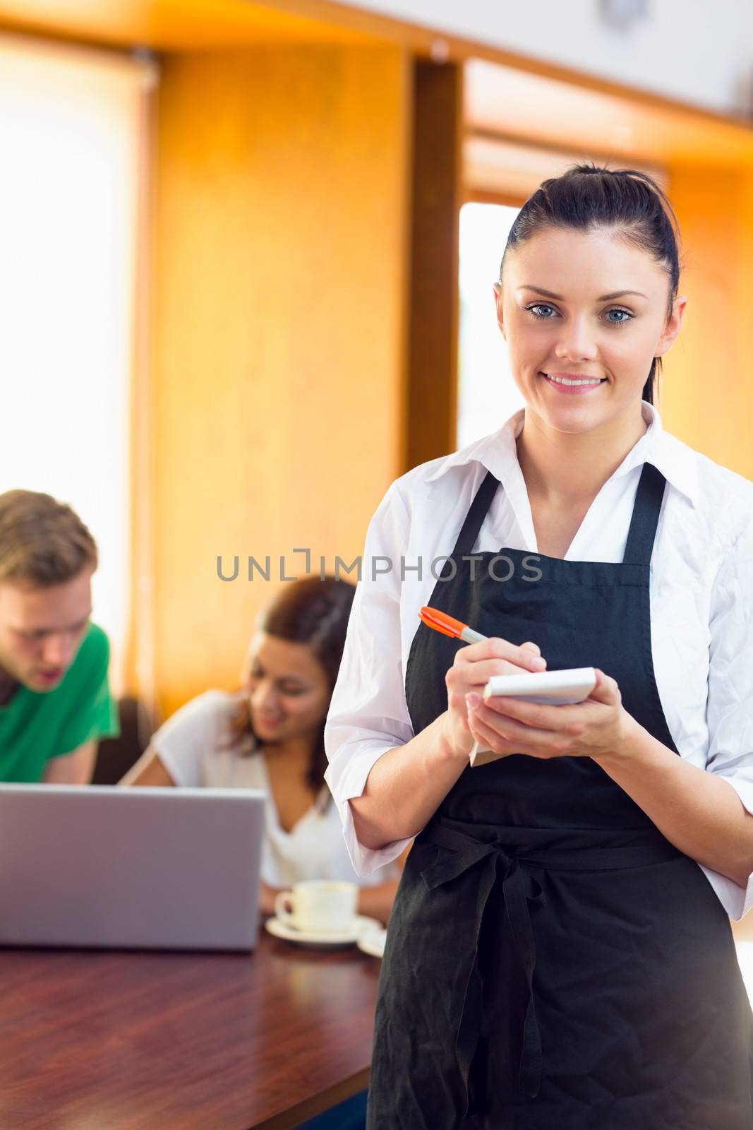 Portrait of a beautiful waitress writing an order with two students using laptop at  the coffee shop