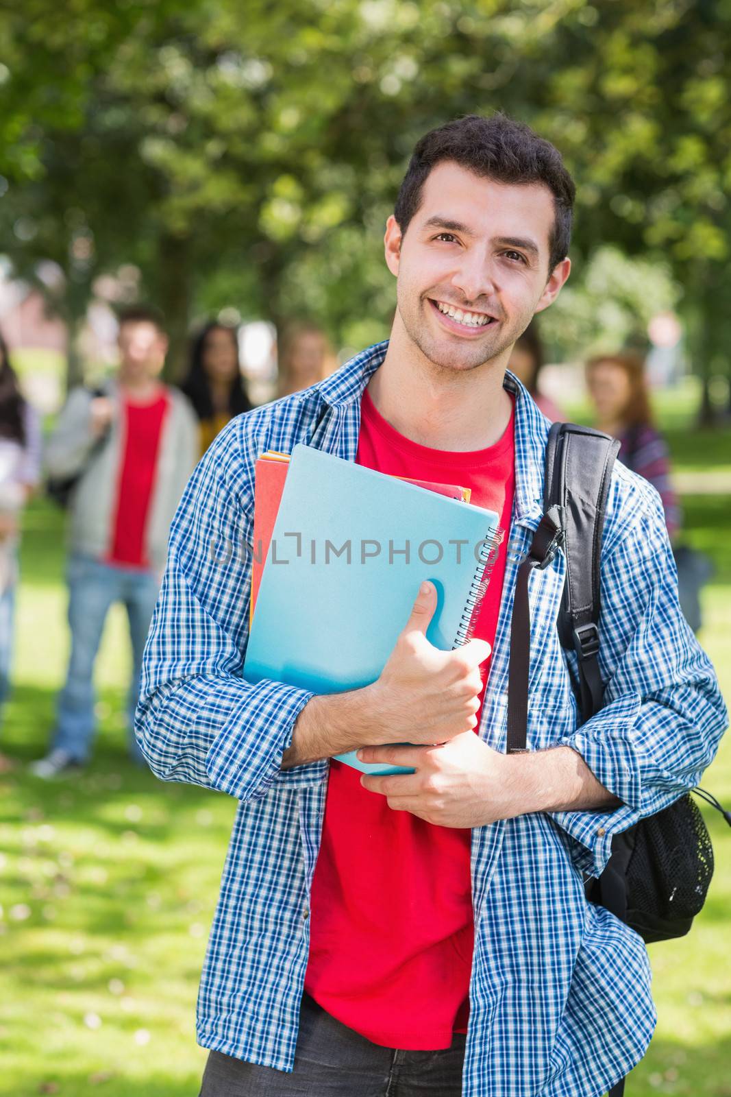 Portrait of college boy holding books with blurred students standing in the park