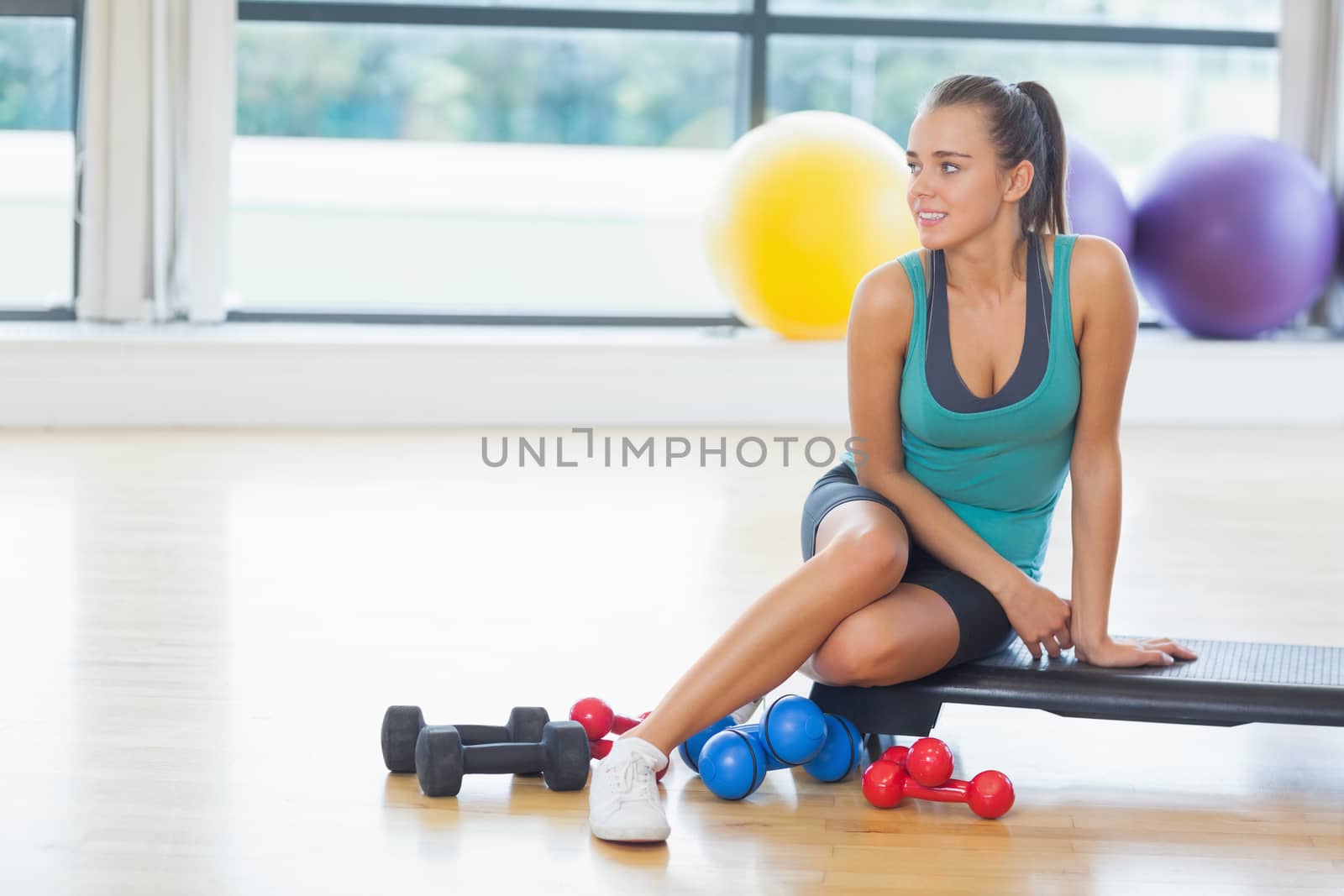 Full length of a young woman sitting with dumbbells in fitness studio