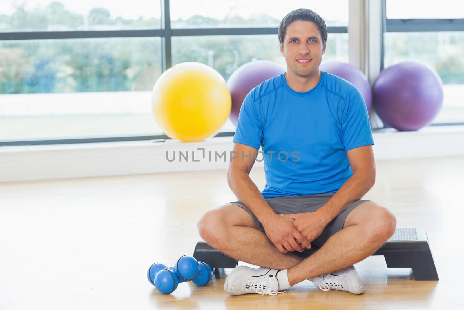 Full length portrait of a young man sitting with dumbbells in fitness studio