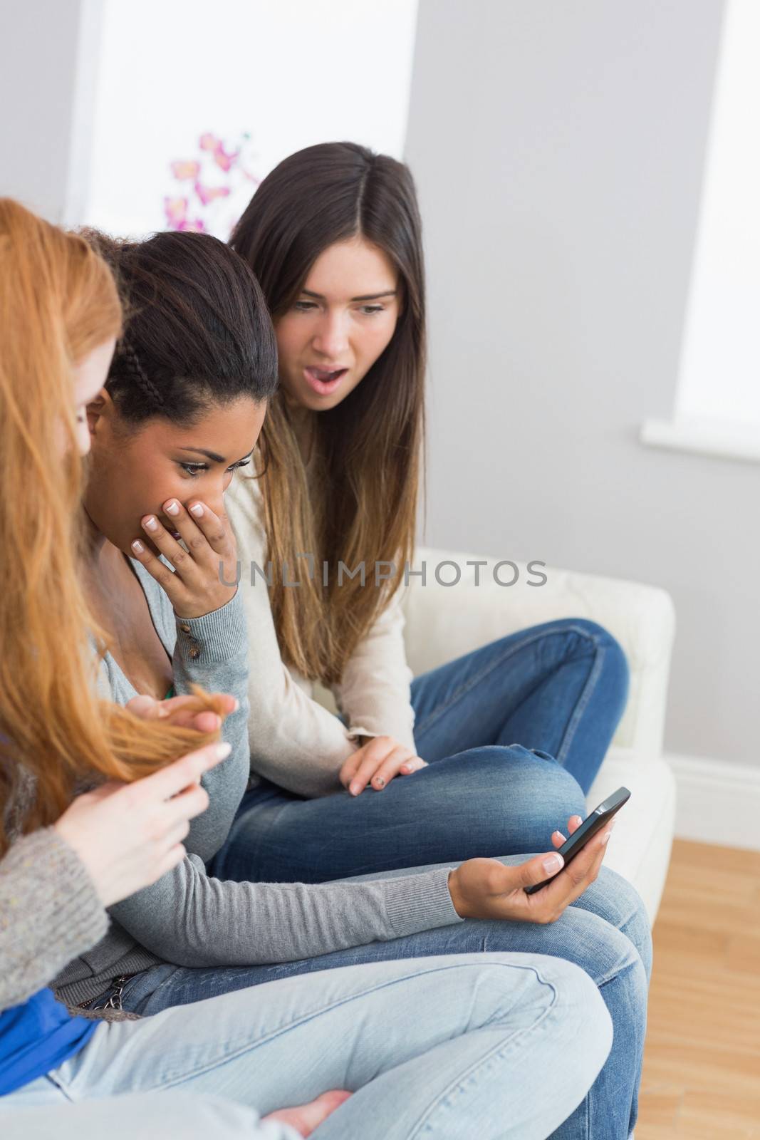 Shocked young female friends looking at mobile phone together on sofa at home