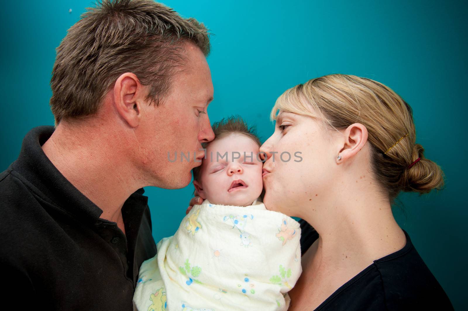 Young father and mother kissing sweetly their beautiful and lovely newborn little daughter isolated on a blue background