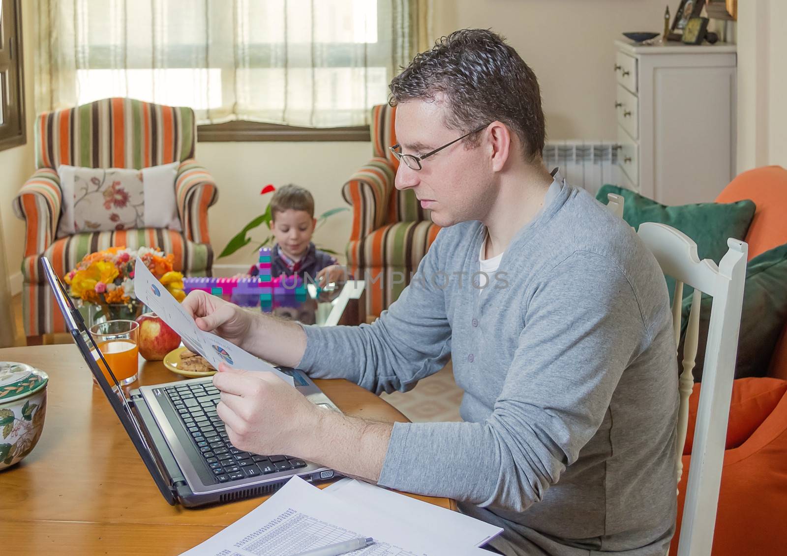 Father reading business documents in home office and his boring son playing on the background