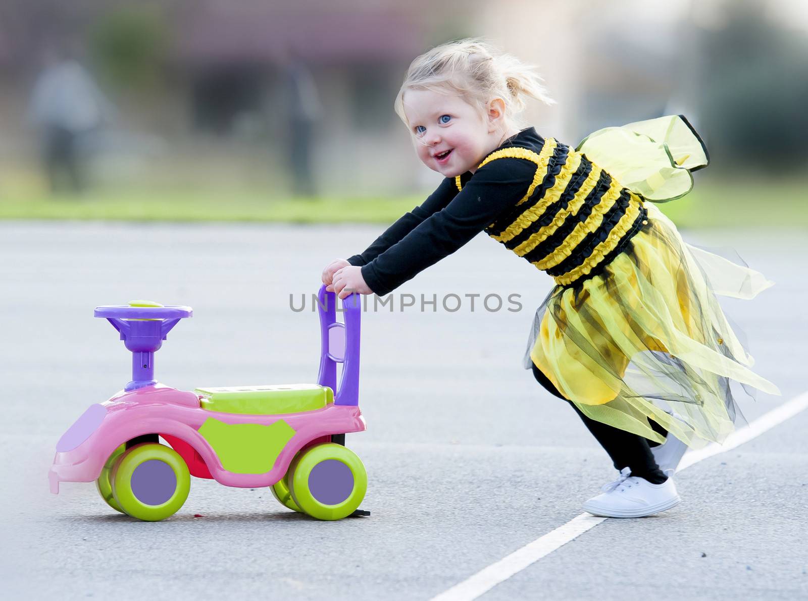 Happy blond little girl in bee costume pushing toy outdoors