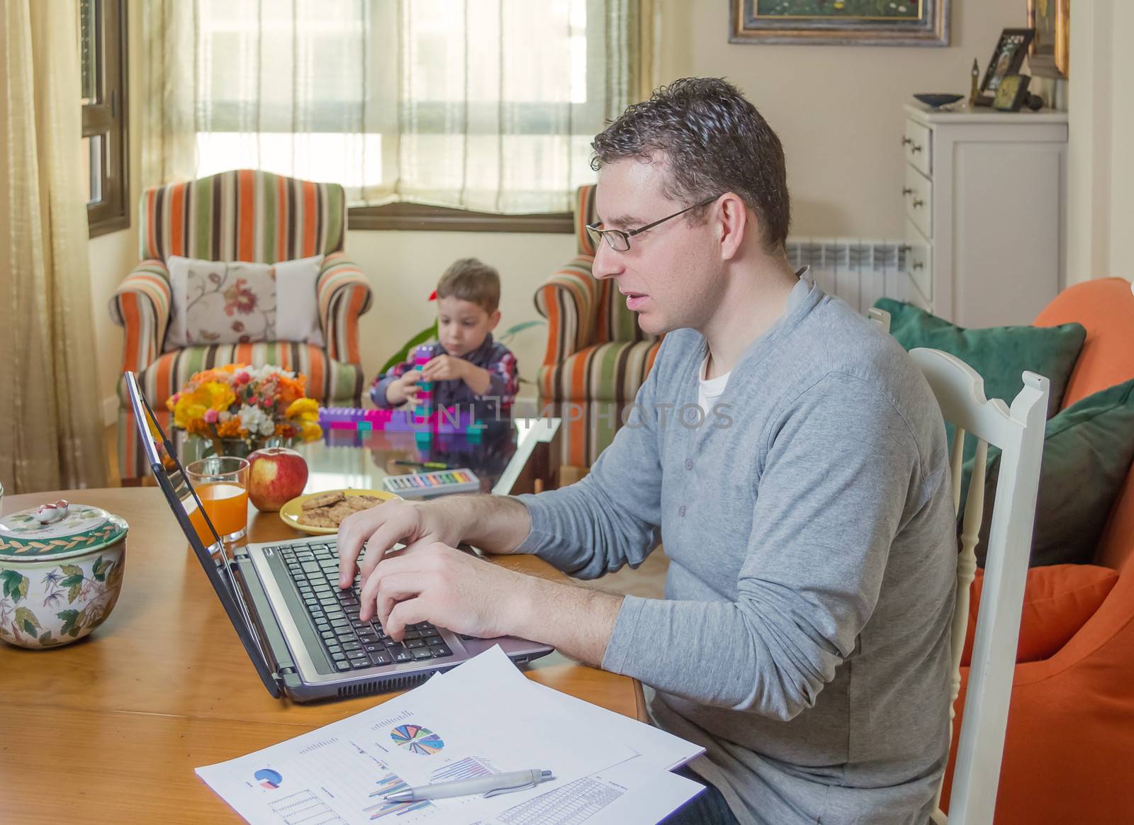 Father working hard in home office with notebook and his boring son playing on the background