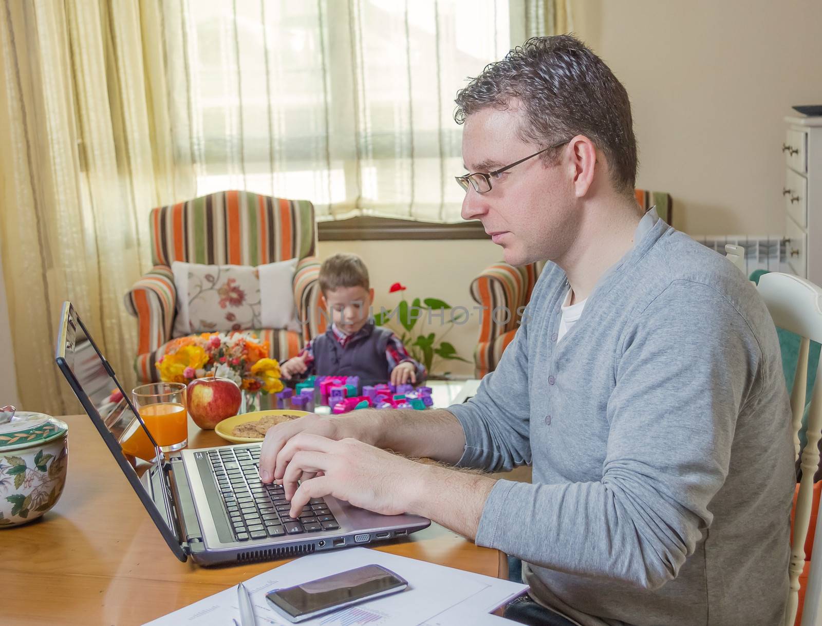 Father working hard in home office with notebook and his boring son playing on the background