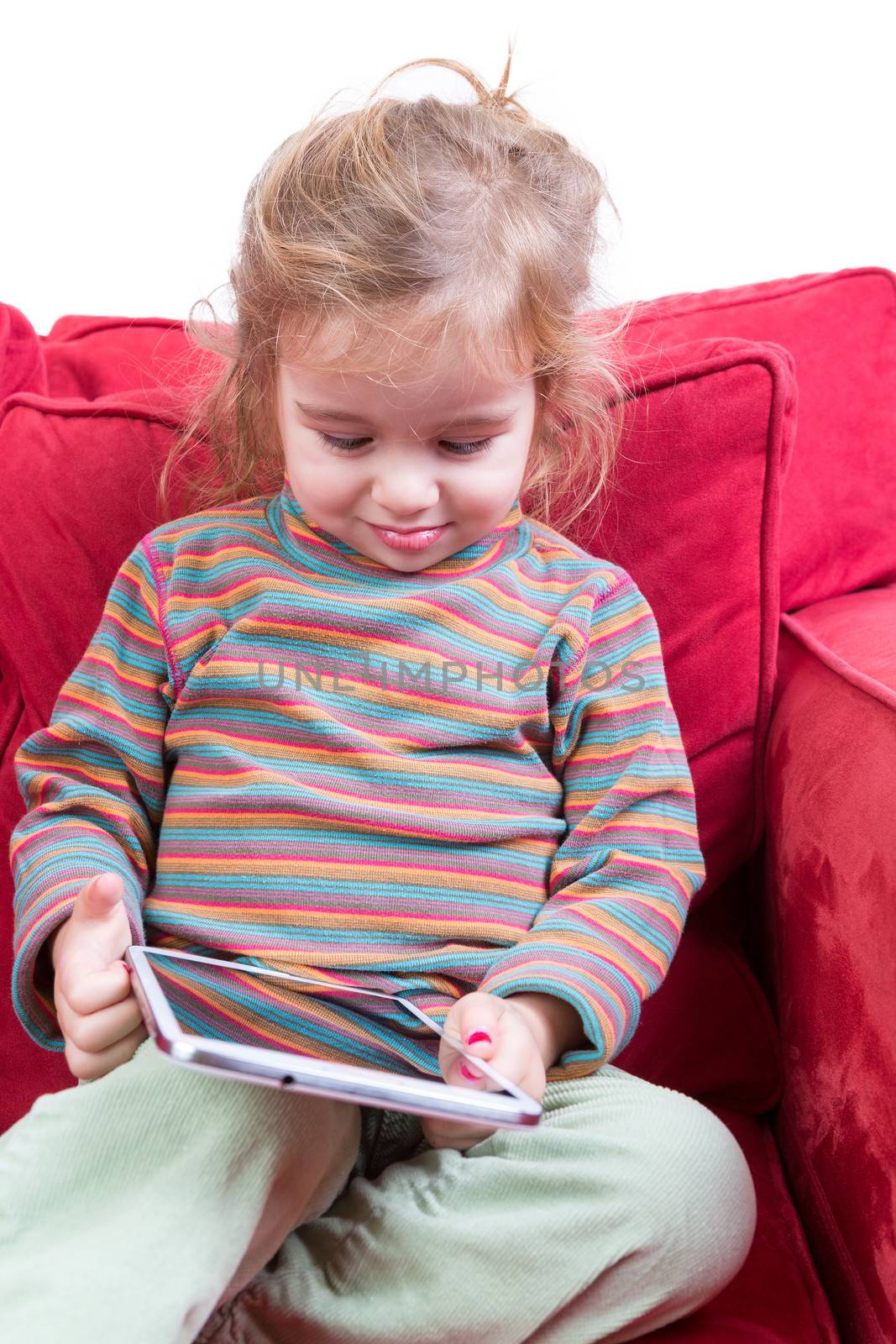 Adorable shy little girl with a tablet-pc in her hands sitting smiling with downcast eyes on a red sofa