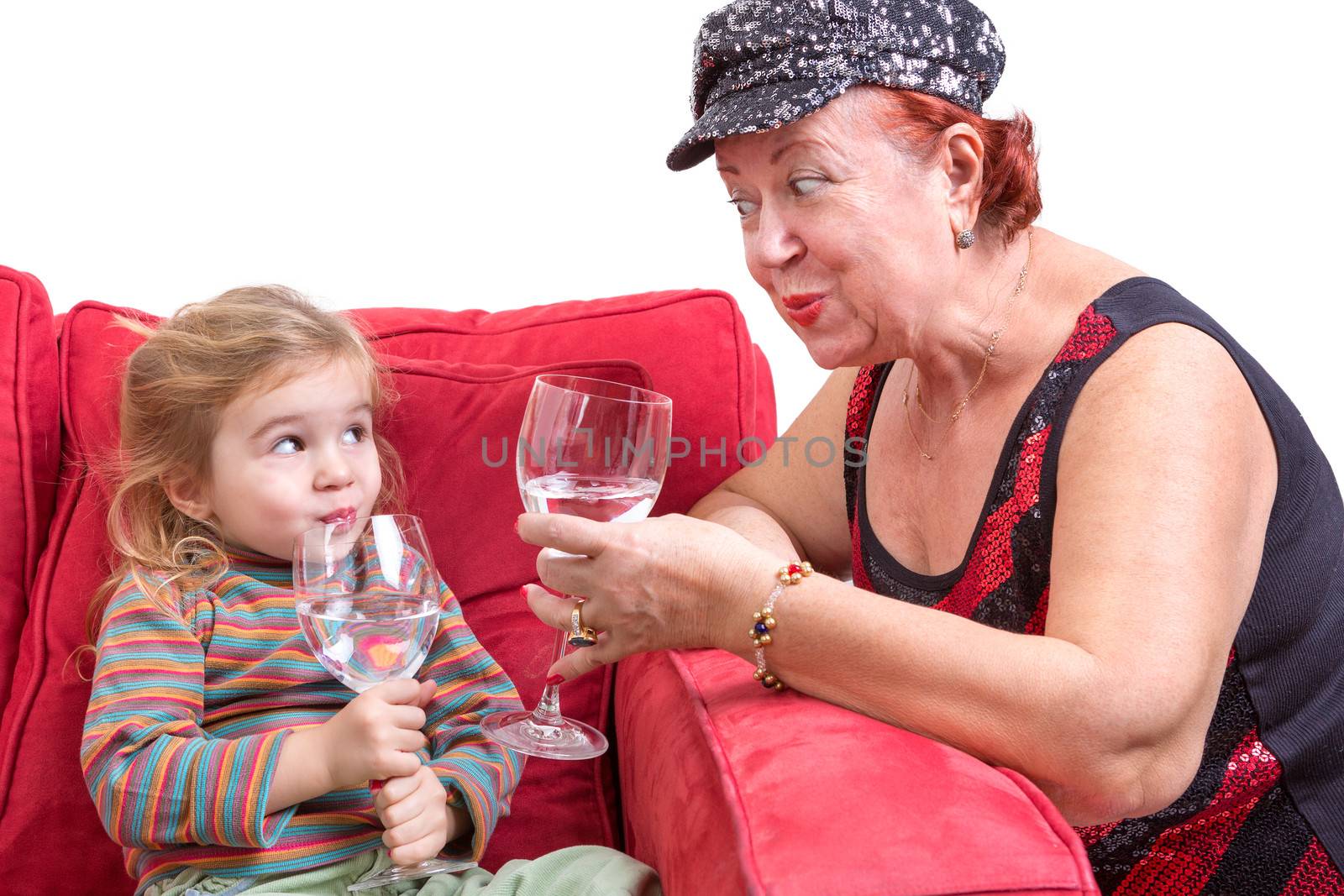 Mischievous trendy grandmother and her beautiful little granddaughter toasting each other with wineglasses of chilled water as the little girl snuggles down in a comfortable red sofa