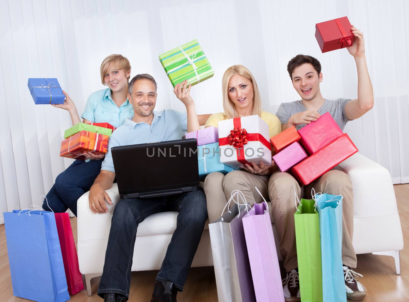 Happy family of online shoppers laden with gifts sitting around the father who is sitting with his laptop computer on his lap