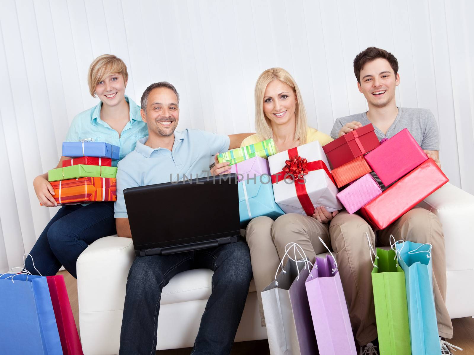 Happy family of online shoppers laden with gifts sitting around the father who is sitting with his laptop computer on his lap