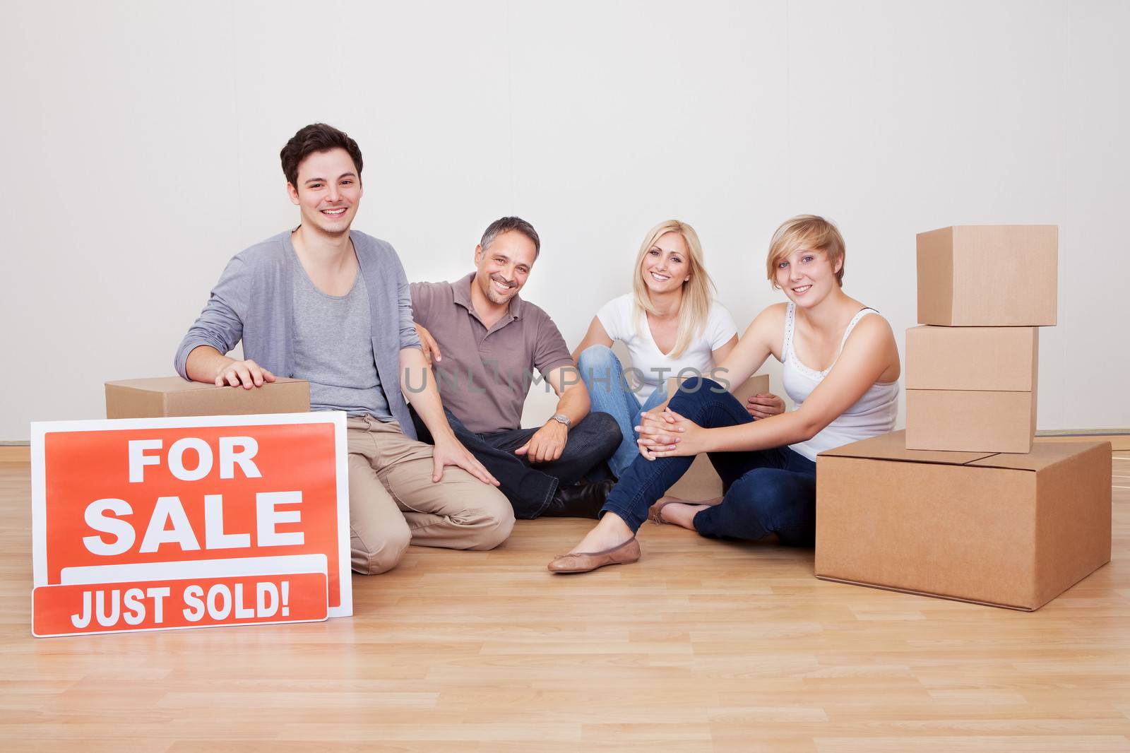 Happy family packing up their home sitting in an empty room on the floor with a sold sign and a small stack of cardboard cartons