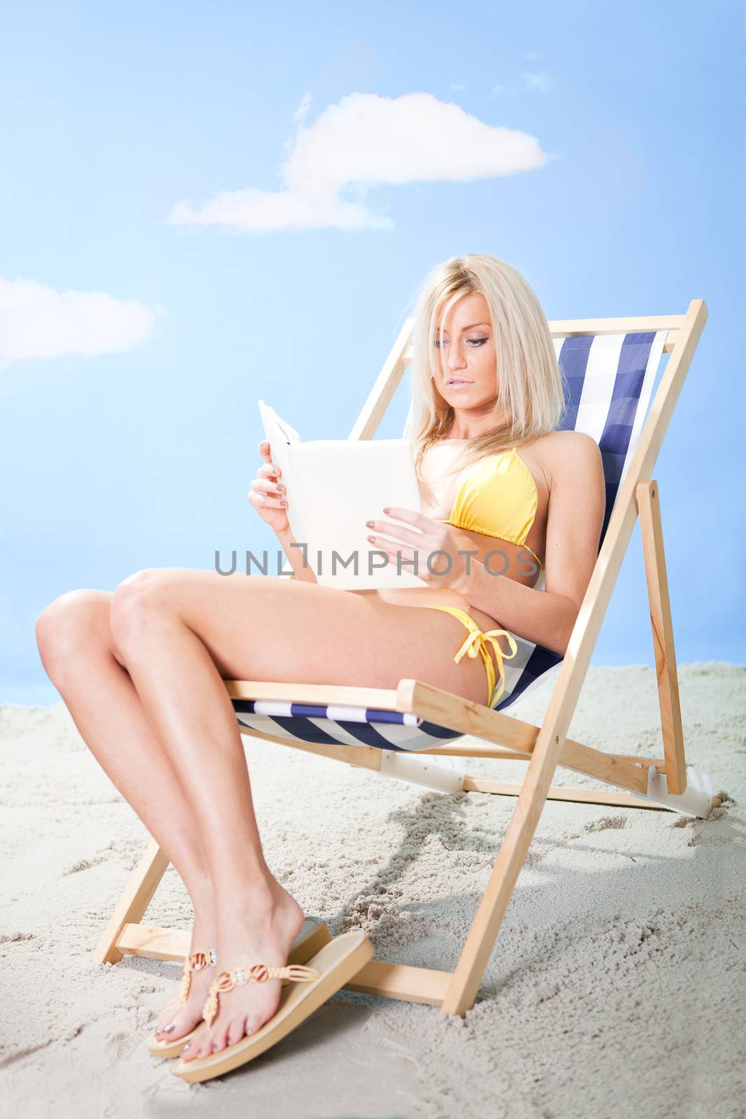 Beautiful young woman in bikini lying on a deckchair and reading a book at the beach