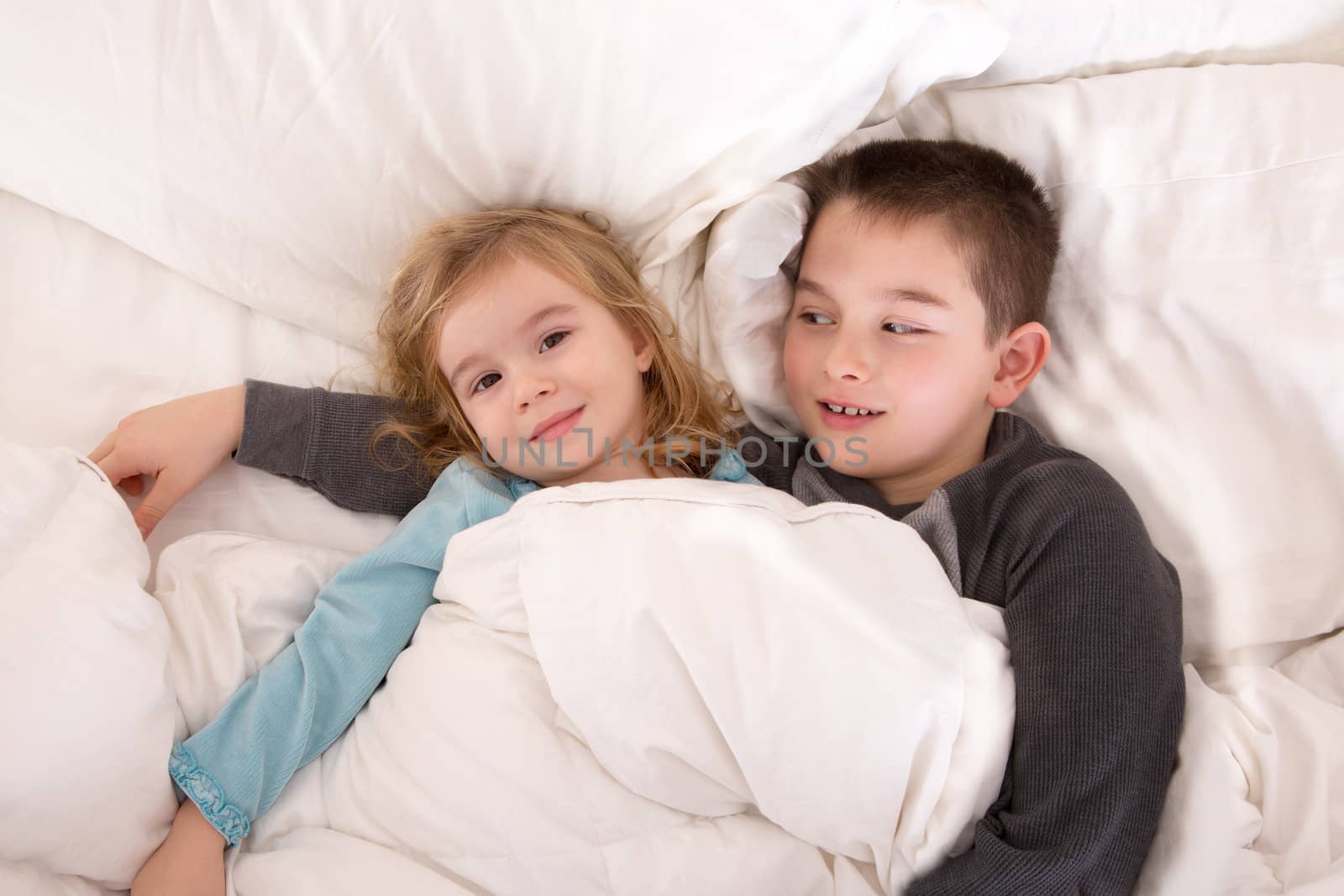 Protective young boy lying in bed with his little sister watching over her as she lies smiling up at the camera as they prepare to go to sleep